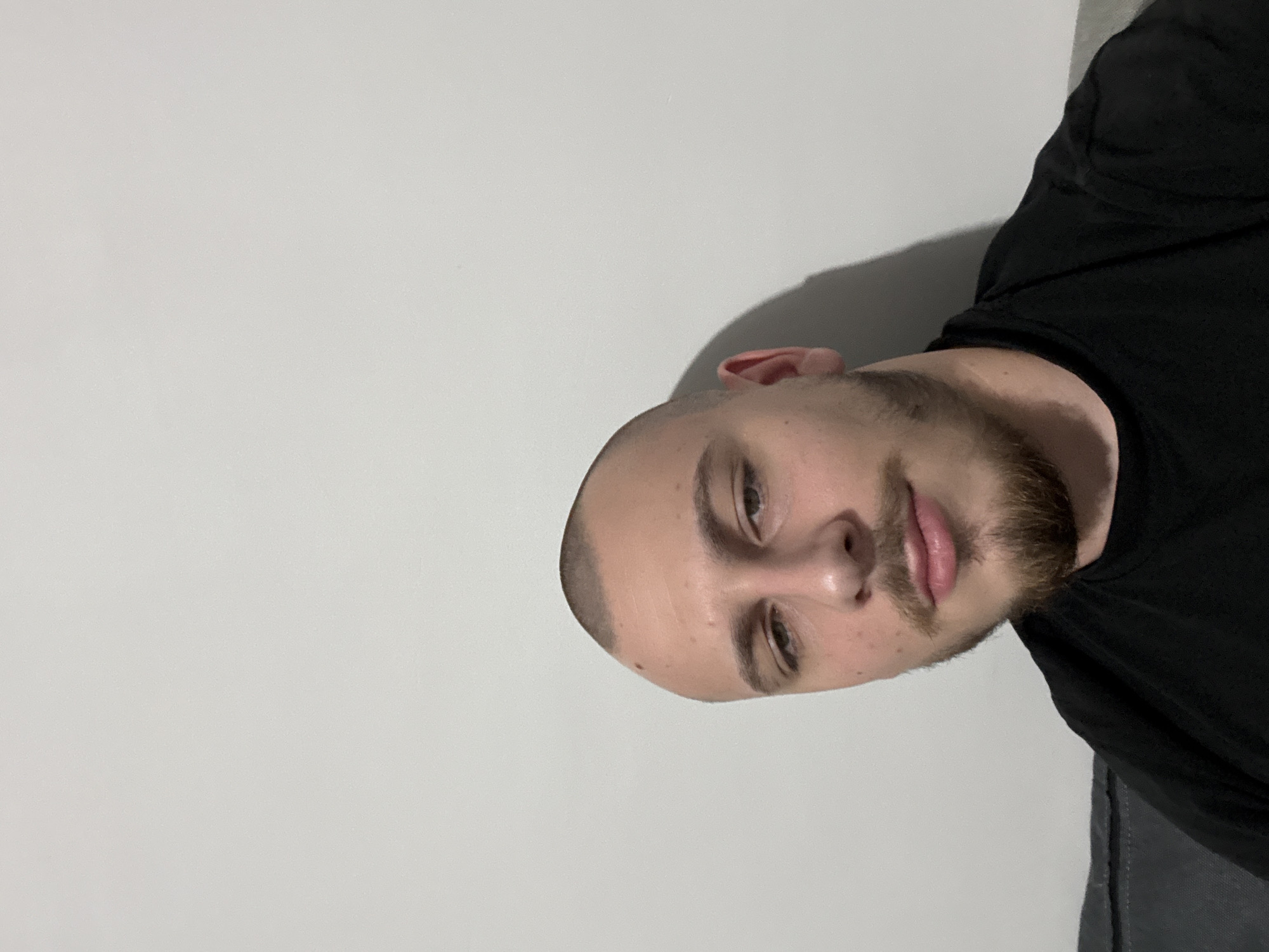 A man with closely cropped hair and a beard is positioned in the lower right quadrant of the image, wearing a black shirt. A large, off-white wall occupies the majority of the frame to the left and above him. The location is not discernible from the image.