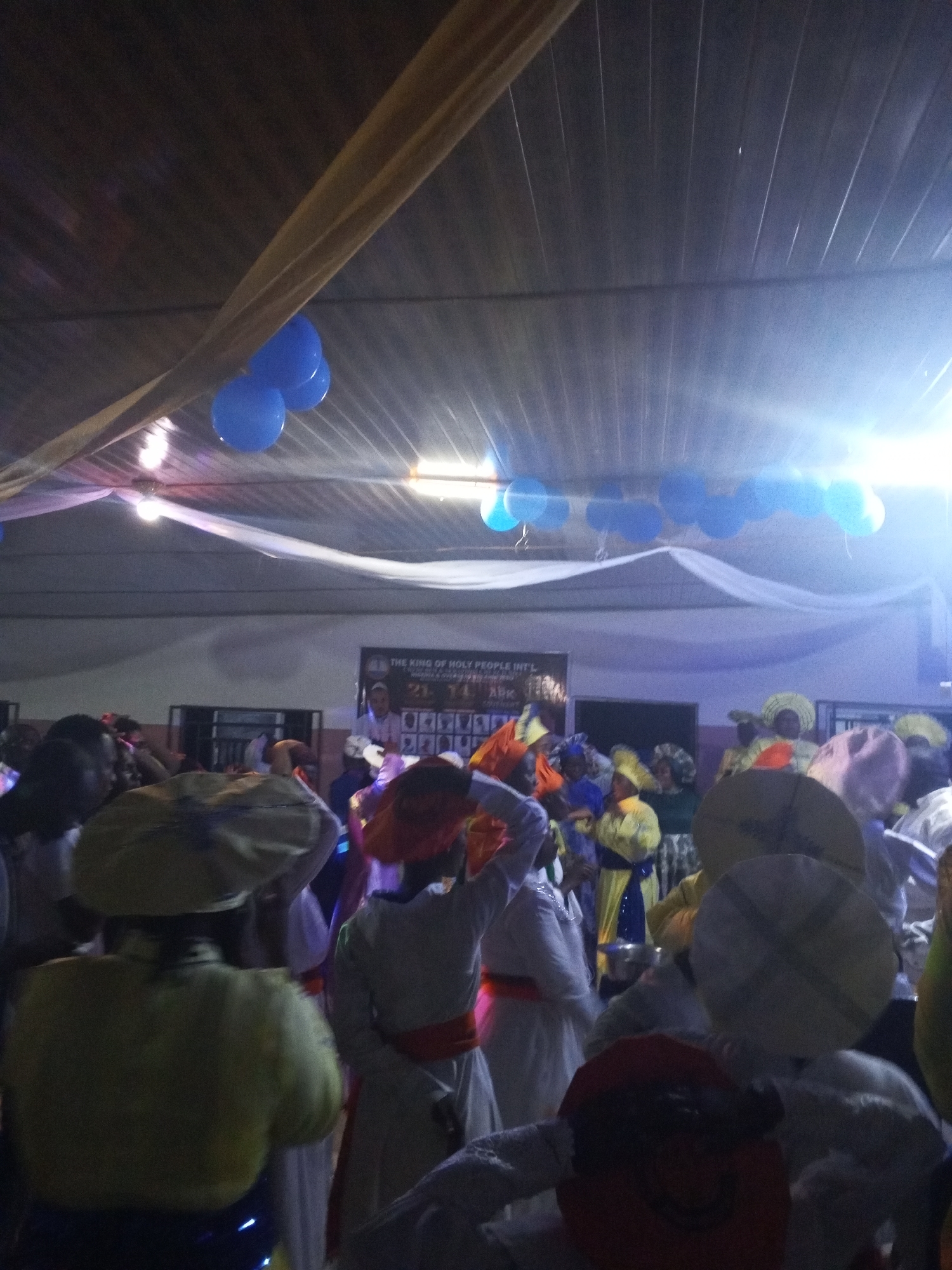 The image shows a gathering of people inside a hall in Lokoja, Nigeria. Many are wearing similar white gowns with colored sashes and head coverings. Blue balloons and white drapery are hung from the ceiling. A banner reading "THE KING OF HOLY PEOPLE INT'L" is visible in the background with a photo of a man.