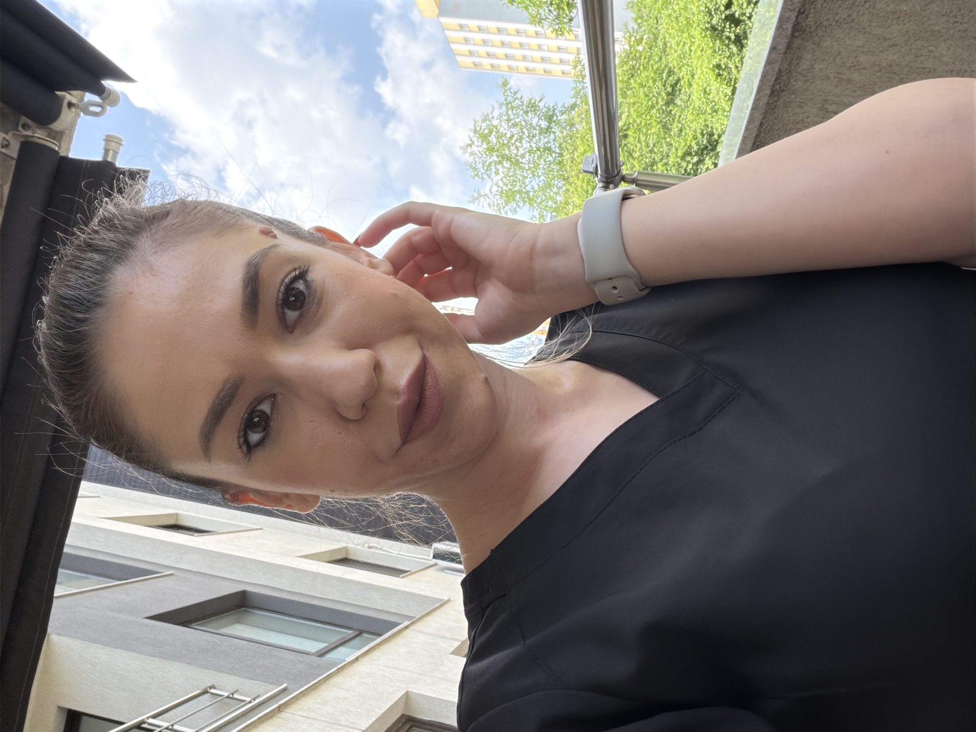 The image captures an adult female taking a selfie-style photograph outdoors, in the city of Sofia, Bulgaria. The subject, viewed from the upper chest to the top of her head, has fair skin, dark eyes, and dark, prominent eyebrows. A small, raised mark is present on her forehead above her left eye (viewer's left). Her dark hair is styled back, with fine strands visible near her temples and ears. She wears a black, V-neck uniform top, appearing to be a scrub. Her lips are full and colored with a muted brownish-mauve lipstick. Her left hand is raised, with fingers touching her left ear, showcasing a light grey smartwatch on her left wrist.
The immediate background above the subject reveals a dark, retractable awning or canopy structure, including its metal fixtures. To the left, a portion of a modern, multi-story building with a light-colored facade, dark-framed windows, and balconies with metal railings is visible. In the mid-ground to the right, lush green trees with dense foliage are present. Further in the distance, a tall, multi-story building with a distinct yellow and white facade and a grid pattern of windows can be seen. The sky is blue with scattered white clouds, indicating daylight and fair weather conditions.