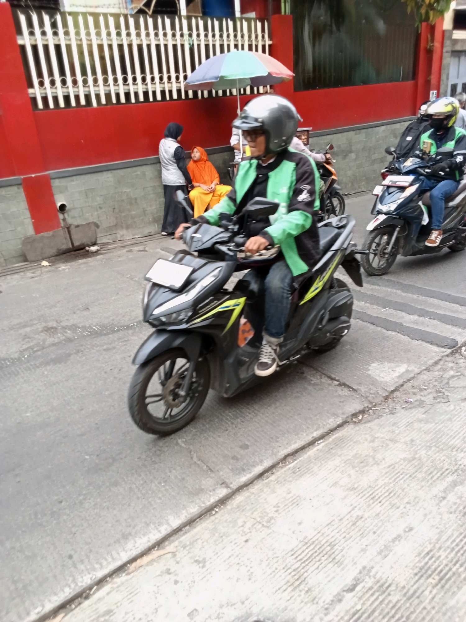 A male motorcyclist wearing a green and black jacket, blue jeans, and a gray helmet is riding a black scooter. He is on a street with other people and scooters in the background. The scooter has a license plate that is not legible. In the background, there are buildings with red and white walls, and a white fence. There is also a multi-colored umbrella on the street. The scene appears to be a street in a city.