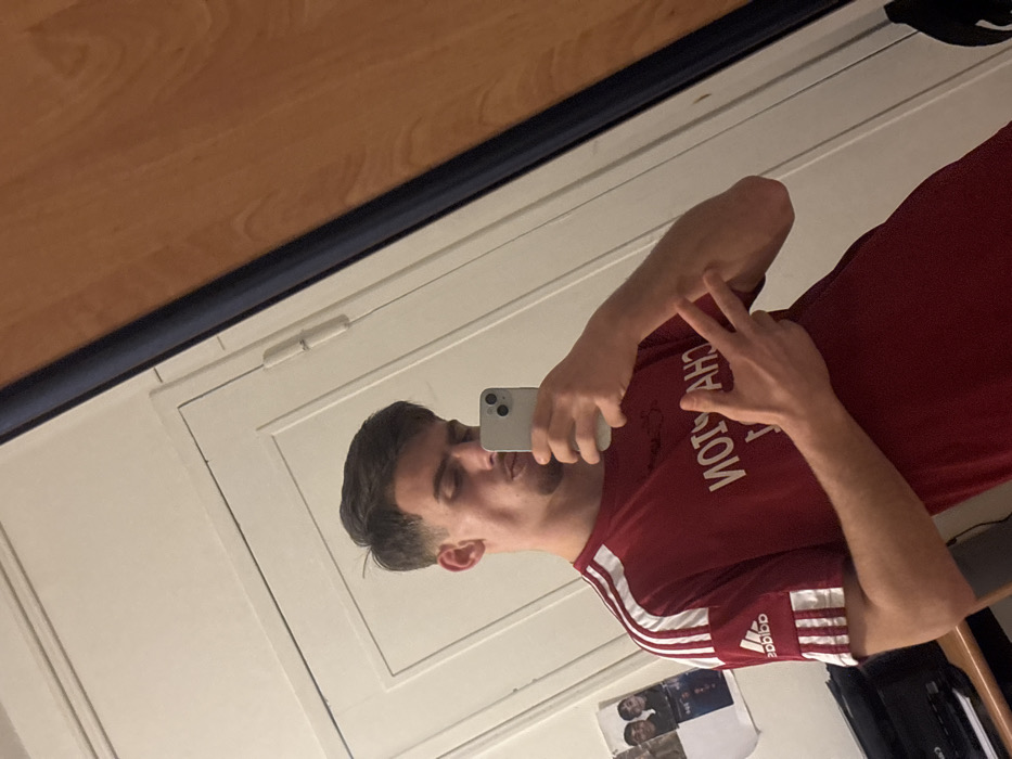 A young man wearing a red Adidas shirt is taking a selfie in a mirror. He is holding his phone in his left hand and making a peace sign with his right. The word “CHAMPION” is printed on the shirt in white letters, which appear reversed in the reflection. A wooden desk and white door are seen in the periphery of the mirror.