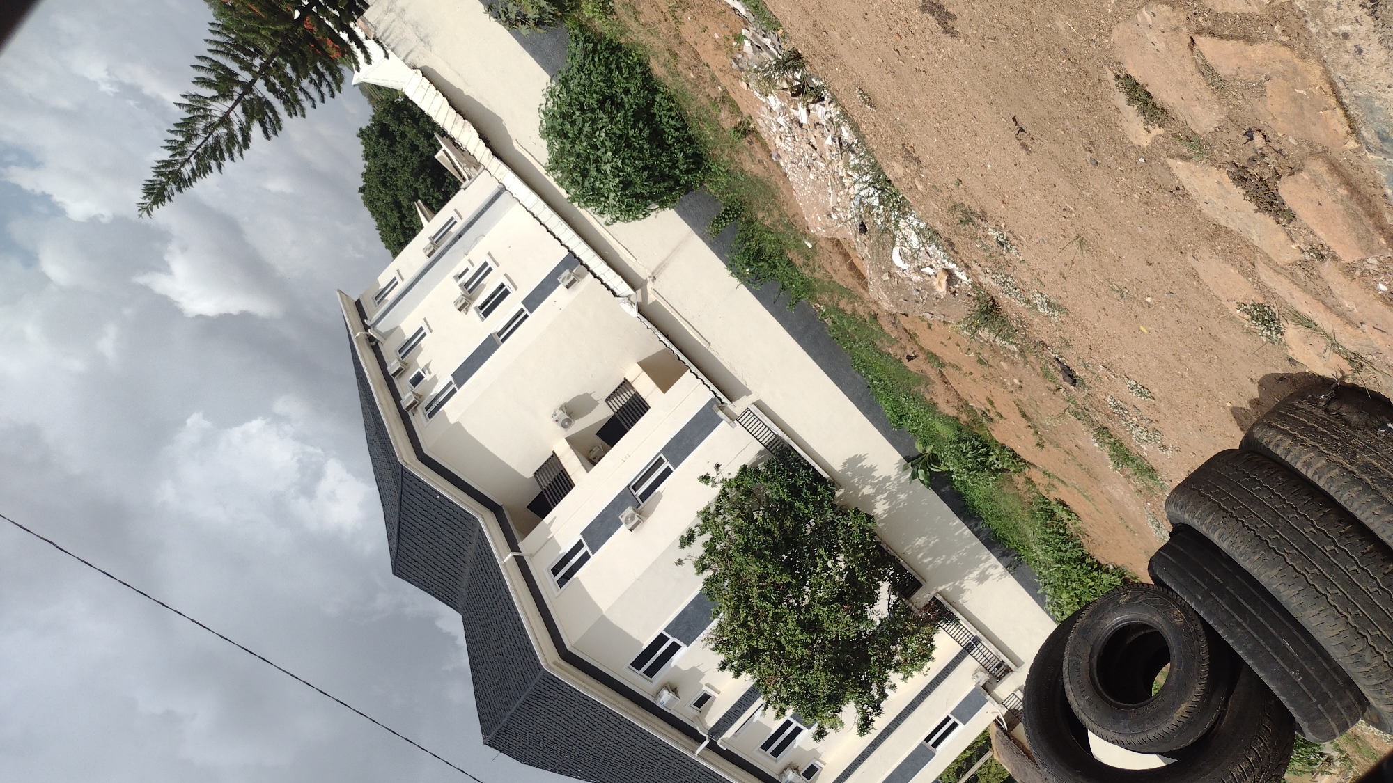 The image depicts a modern multi-story building with a dark gray roof and light-colored walls, accented by darker gray vertical stripes and numerous windows. Multiple air conditioning units are affixed to the exterior walls. A large tree is situated in front of the building, partially obscuring the view of some of the windows.
The environment includes an unpaved area with dirt, some vegetation, and debris adjacent to the building's paved walkway. A stack of used tires is visible in the foreground. The sky is overcast with gray clouds. Based on available information, the location is likely in Jos, Nigeria.