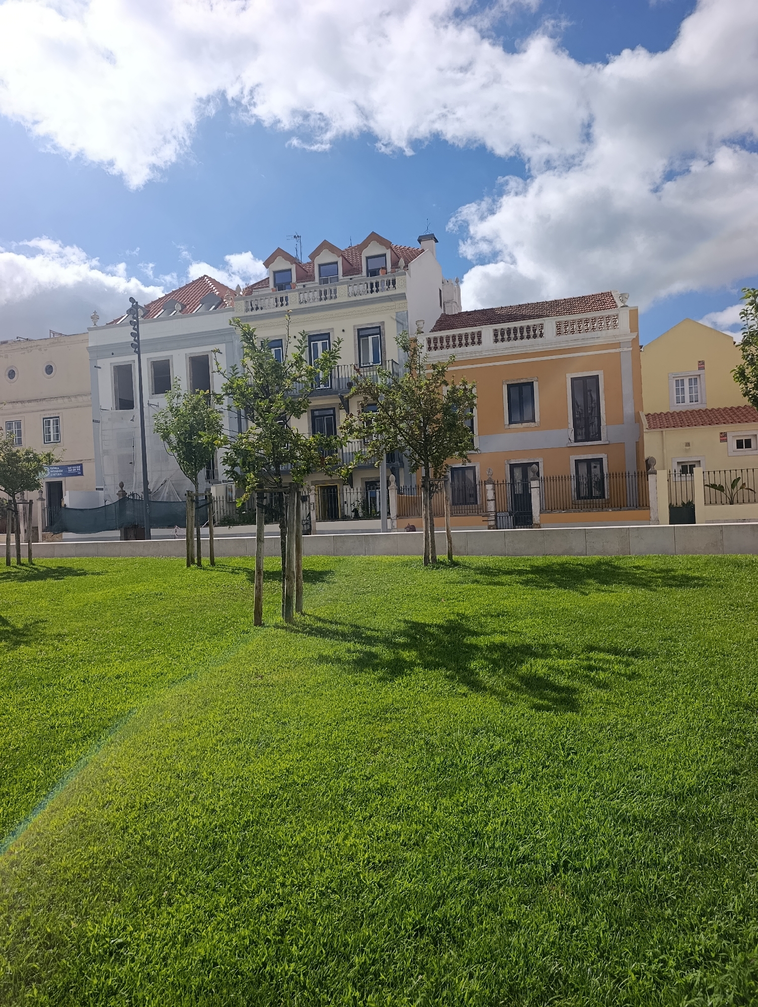 This is a photograph of a modern urban park area with buildings in the background. In the foreground, a meticulously manicured green lawn occupies the majority of the frame. Sunlight creates lens flare on the left side of the lawn, and shadows from trees are cast across the grass. Several young trees, supported by wooden stakes, are planted in rows on the lawn.
Behind the park, a row of multi-story buildings with distinct architectural styles lines the street. The buildings feature various facade colors, including pale yellow, light blue-grey, and a prominent orange structure with white trim. Several windows and balconies are visible. One building on the left appears to be under renovation, with scaffolding and netting.
In the sky above the buildings, bright white clouds are scattered across a clear blue sky. The scene depicts a well-maintained public space in a city, likely a sunny day. While the specific location within Setúbal, Portugal, is not identifiable from visual cues alone, the architecture suggests a European setting. There are no people or animals visible in the image.