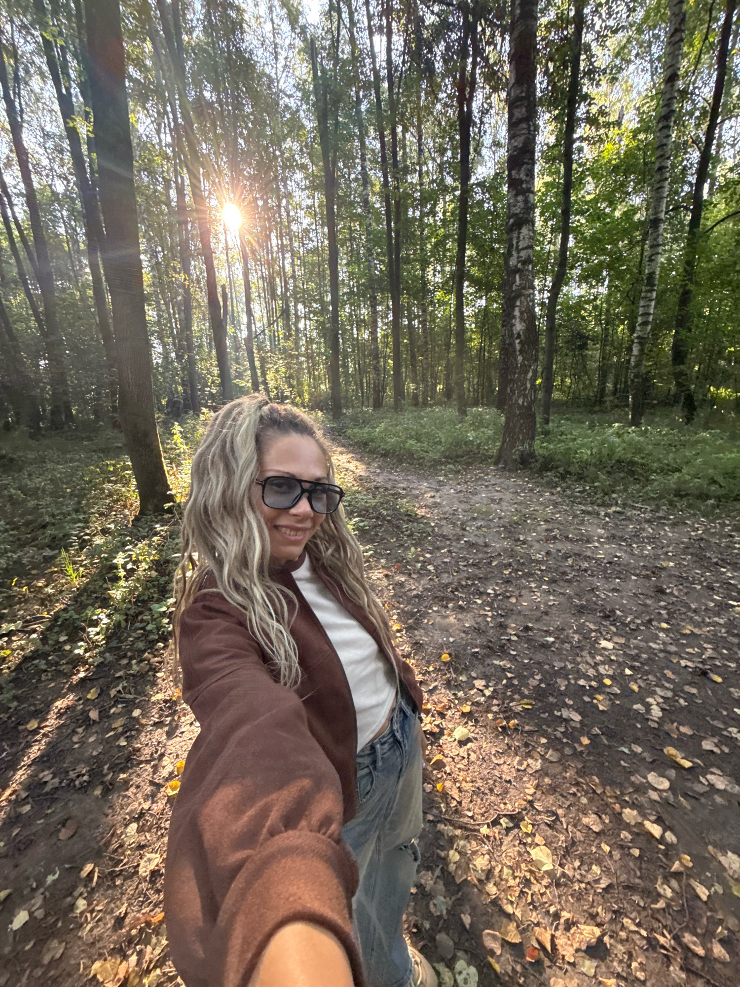 A blonde woman with curly hair is taking a selfie in a forest in Rusanovo, Russia. She is wearing sunglasses, a brown jacket over a white top, and ripped jeans. The sun is shining through the trees, casting long shadows on the leaf-covered ground. The trees are tall and thin, with some bare branches and others covered in green leaves. The ground is covered in fallen leaves, twigs, and some patches of green vegetation. The overall atmosphere is peaceful and natural, with a hint of autumn.
