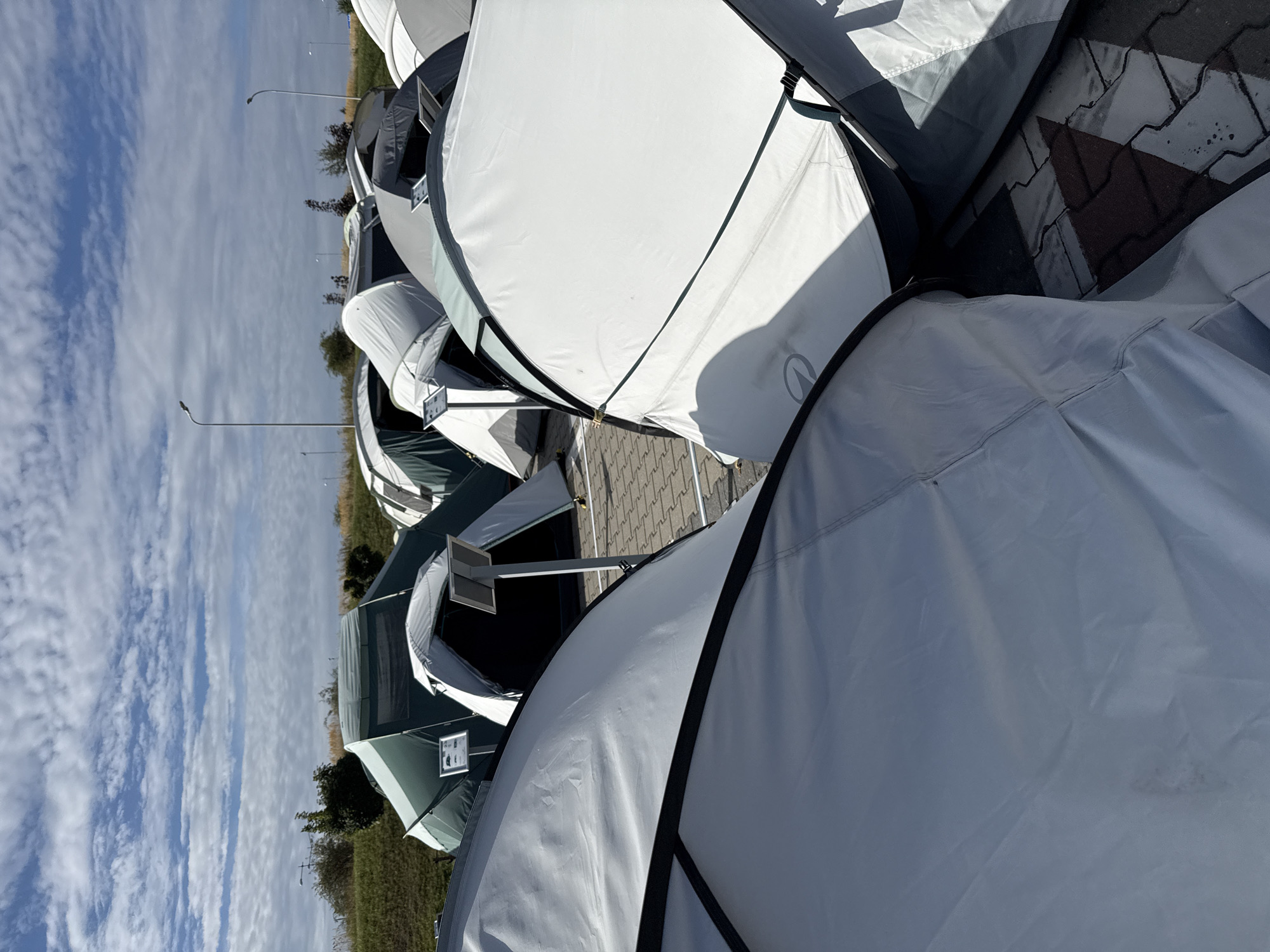 The image shows an outdoor display of multiple tents on a paved area, possibly a parking lot, in Bielany Wrocławskie, Poland. The tents are of various sizes and colors, predominantly white, gray, and green, and appear to be set up for display or sale. Some tents are open, revealing their interior structures, while others are closed. In the background, there is a grassy embankment with scattered trees and utility poles. The sky is partly cloudy with a mix of blue sky and white clouds. The ground is paved with interlocking bricks. There are no people visible in the image.
