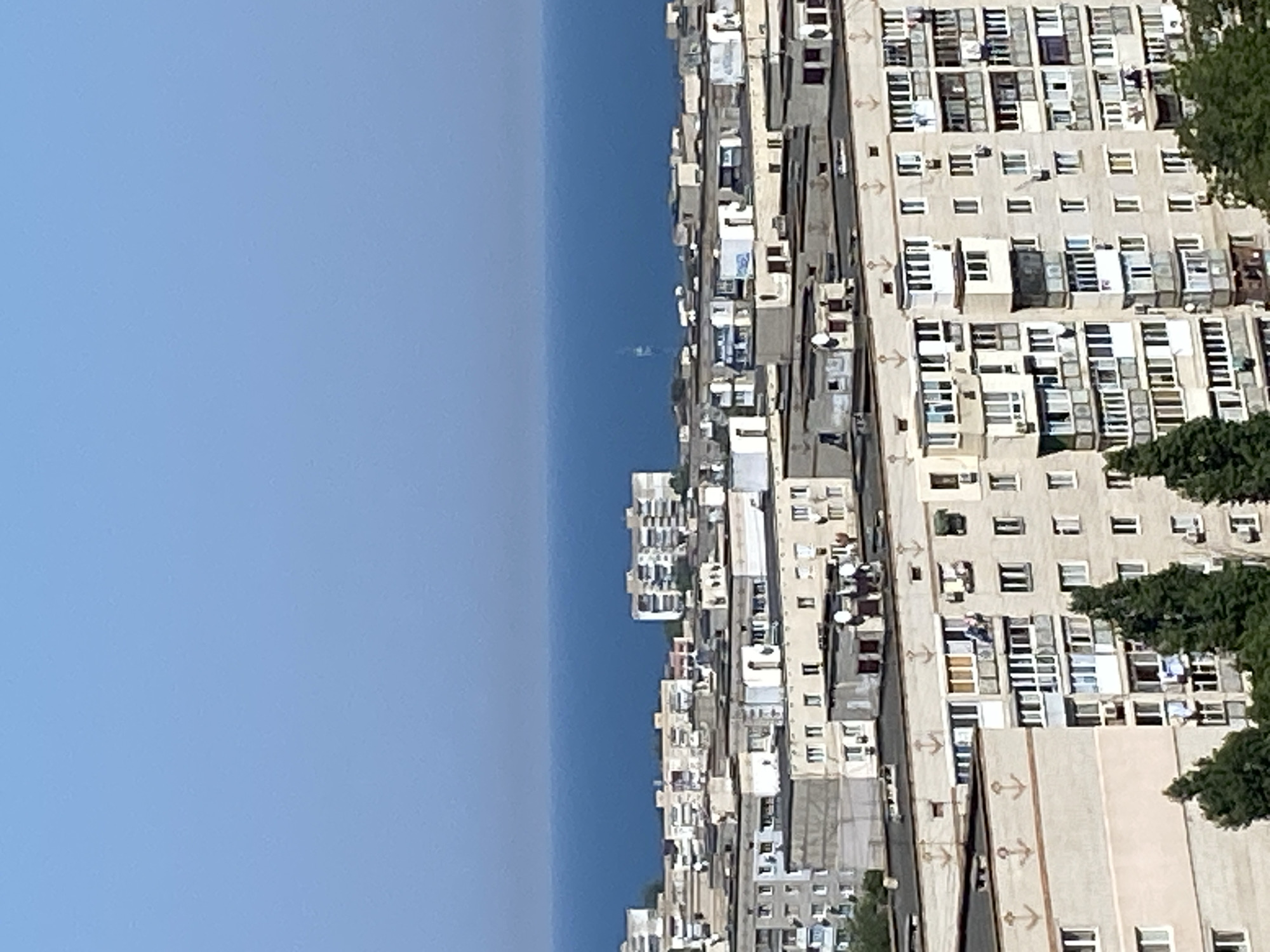 The image depicts an aerial view of an urban landscape in Chornomorsk, Ukraine, overlooking a body of water, likely the Black Sea. The foreground is dominated by a dense cluster of Soviet-era apartment buildings, characterized by their uniform design and concrete facades. Many of these buildings feature rows of windows, balconies, and external air conditioning units. Some of the buildings have decorative anchor motifs painted on their upper levels. Trees are visible at the lower edge of the frame, suggesting some greenery within the urban setting. In the distance, the sea stretches out under a clear blue sky, with a very faint silhouette of a structure or vessel visible on the horizon. The overall impression is one of a densely populated coastal city.