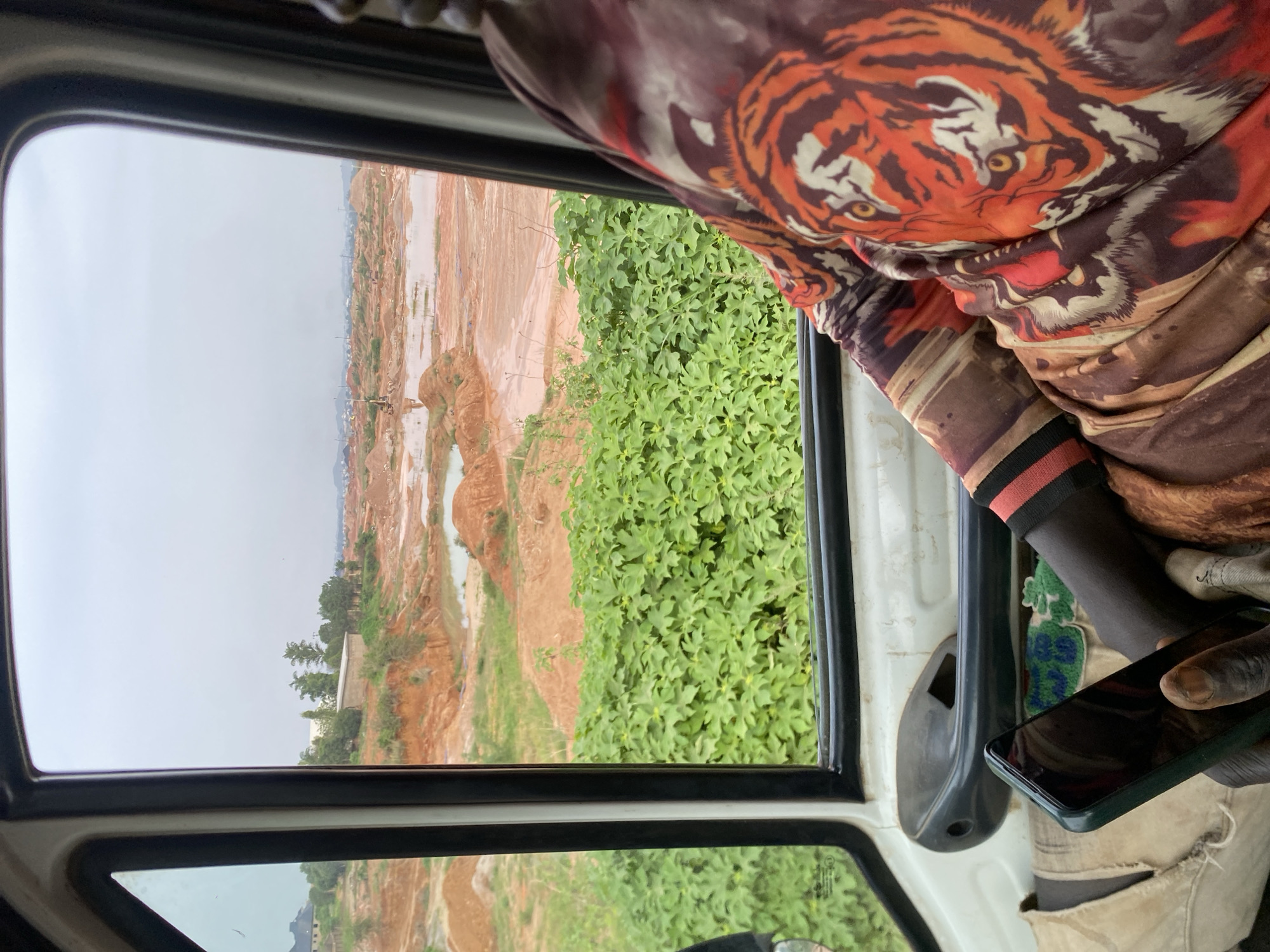 An interior view from the driver's side of a vehicle in Reifild-Ist, Nigeria shows a landscape of a red clay quarry under a cloudy sky. The quarry is characterized by mounds of reddish-brown earth and several pools of water. Dense green foliage borders the quarry in the mid-ground. In the foreground, a person wearing a shirt with a tiger print is visible from the shoulder down, holding a smartphone. The person's left arm rests on the vehicle's interior, which includes a door handle and part of the seat. The overall scene depicts an active or recently active construction or mining site.