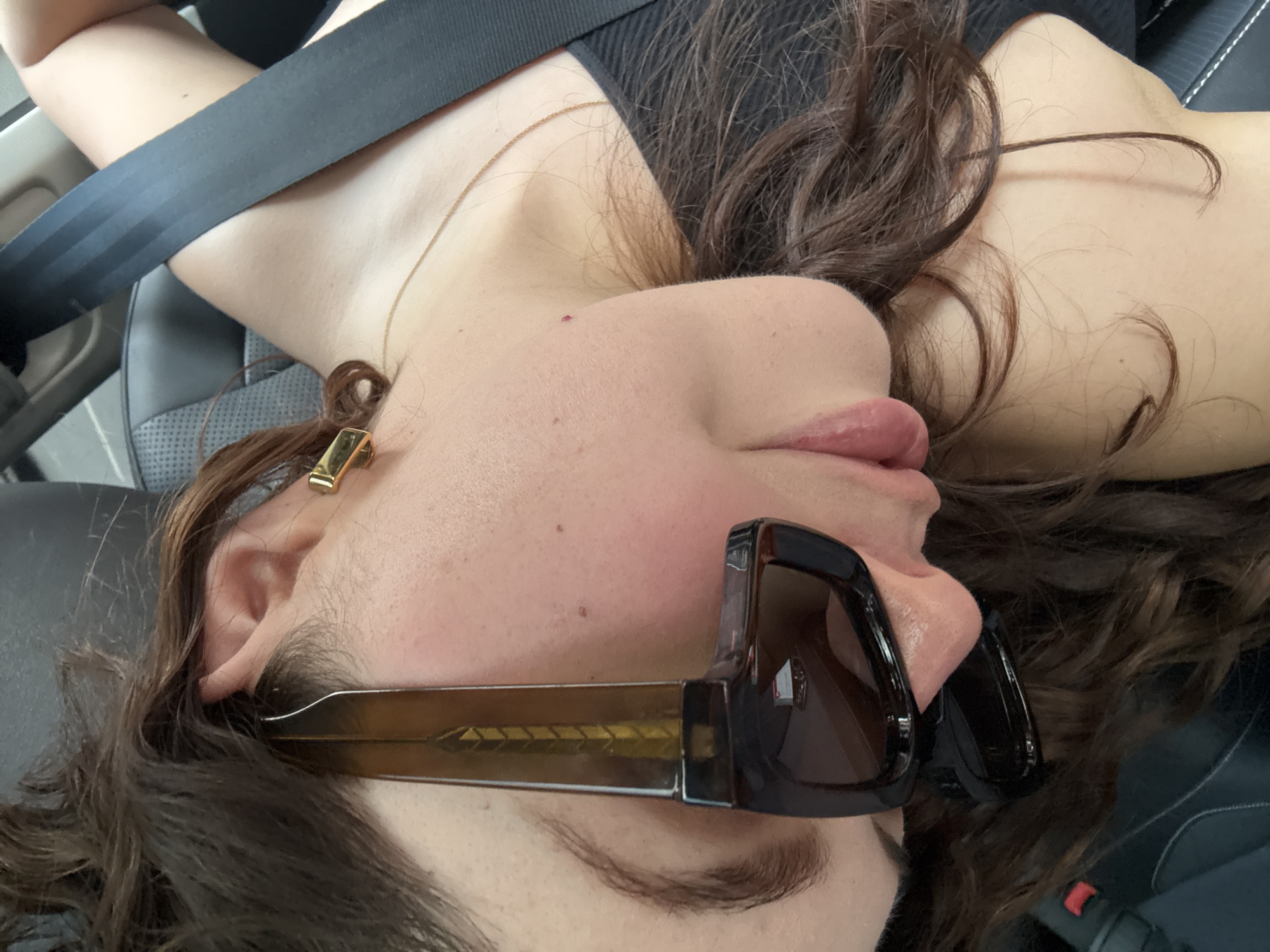 The image presents a close-up, angled view of a person reclining inside a car, likely in Mexico City, Mexico. The subject, possibly female, has fair skin, full lips, and long, wavy dark brown hair that cascades around their head and shoulders. Their face is shown in profile, looking slightly upwards, with their eyes obscured by large, dark-lensed sunglasses featuring brown/amber frames.
A thin gold chain necklace is visible around their neck and a distinctive gold-colored, rectangular earring hangs from their right earlobe. They are wearing a dark-colored top, possibly a tank top or camisole, part of which is visible over their shoulder. A black seatbelt diagonally crosses their torso and shoulder, indicating they are seated in a vehicle. The car interior features dark, possibly leather, seats with visible stitching. The person appears to be in a relaxed state, perhaps resting or enjoying a journey. There is no visible text in the image.