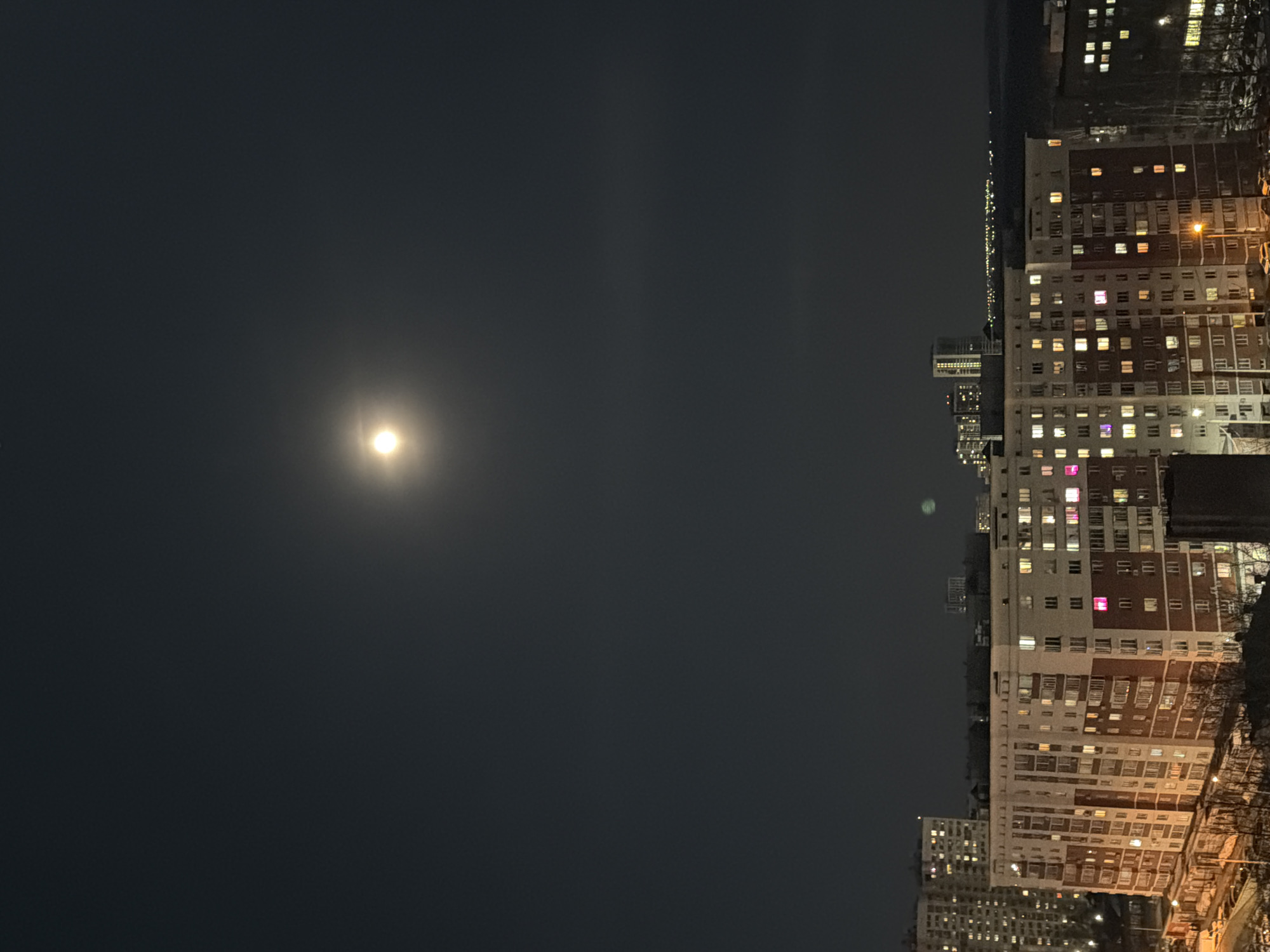 The image depicts a night scene in Izhevsk, Russia, featuring a dark sky illuminated by the moon. In the foreground, a residential area with tall apartment buildings is visible. The buildings appear to be of Soviet-era construction. Lights from individual apartments illuminate the scene, creating a pattern of small rectangles across the building facades. In the distance, the city skyline is marked by other buildings, indicated by their lights. The street lights are visible at the bottom of the image. There are no people visible in the frame.