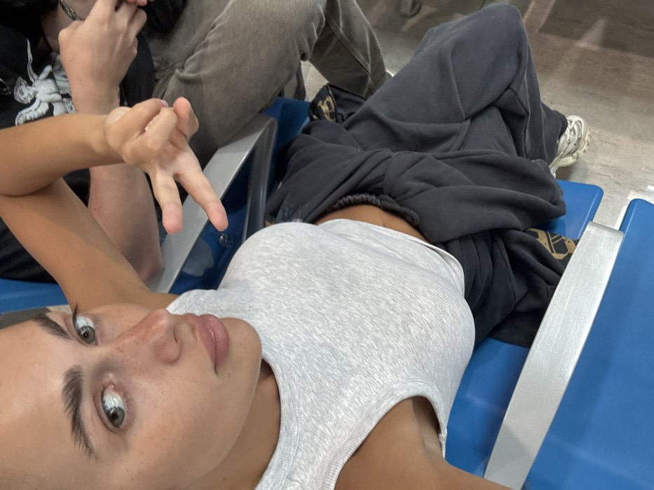 The scene depicts two individuals in a public waiting area, consistent with an airport or ferry terminal in Zanzibar, Tanzania. The primary subject, a person with fair skin, light-colored eyes, prominent eyebrows, and freckles across the nose and cheeks, is positioned in the foreground, looking directly at the camera. This individual wears a light gray ribbed tank top and dark-colored, loose-fitting trousers, with a white athletic shoe visible on the right foot. Their right hand is raised, forming a "V" or peace sign. A second individual is partially visible to the right and behind the primary subject, wearing a dark t-shirt with a white graphic depicting an angelic figure on the upper arm/shoulder area, and light brown or khaki pants. Both are seated on blue public waiting chairs featuring a silver horizontal support bar. The floor appears to be a light-toned, unpatterned surface. No specific identifying markers for Zanzibar are visible within the image.