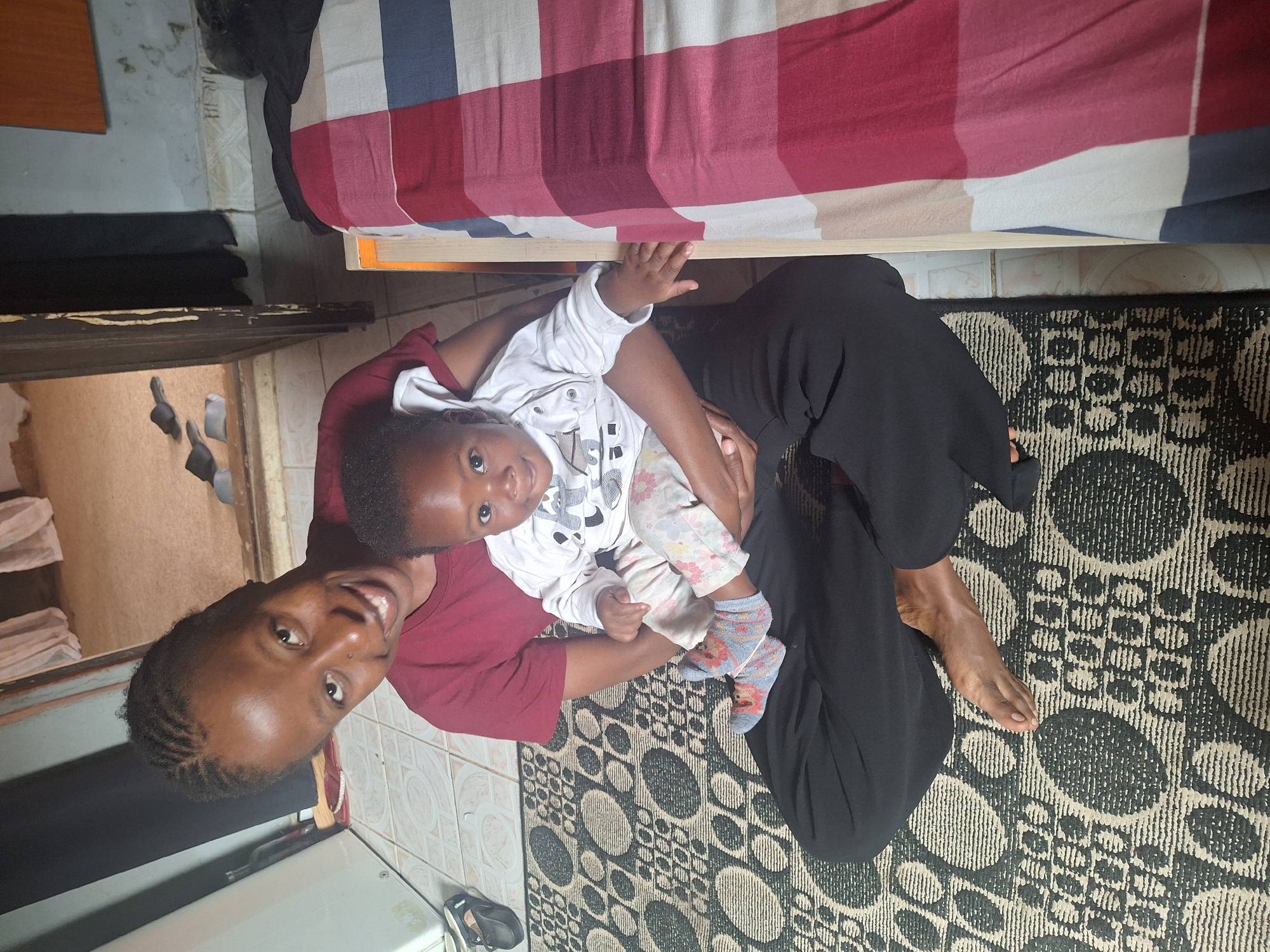 A woman with braided hair is sitting on the floor in Sabon Tasha, Nigeria, holding a baby. The woman is wearing a maroon t-shirt and black pants. The baby is wearing a white long-sleeved shirt with a graphic print and patterned pants. They are sitting on a patterned rug. To the right, a bed with a red, white, and blue plaid comforter is visible. In the background, there are doorways with curtains and what appears to be a closet. Sandals are visible near a white refrigerator-like appliance on the left. The lighting suggests it is daytime.