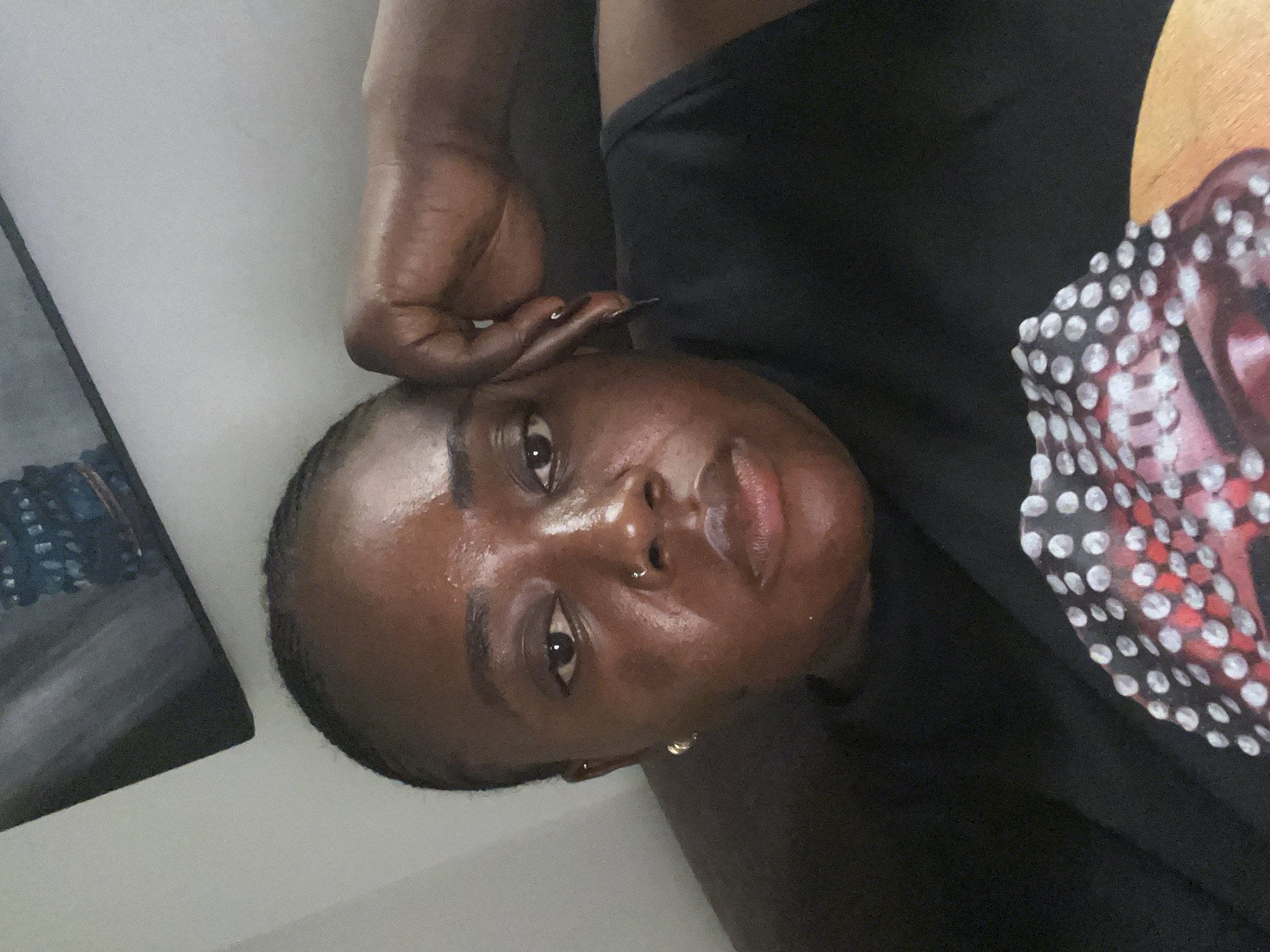A close-up, indoor selfie shows a Black woman with braided hair resting her chin on her hand. She is wearing a black t-shirt with a graphic print of a person's face with red lips and white polka dots. Her left earlobe has a small gold hoop earring. The background is a light gray wall with a portion of a dark-framed picture hanging at an angle in the upper left corner. The scene appears to be a residential setting in Abuja, Nigeria.