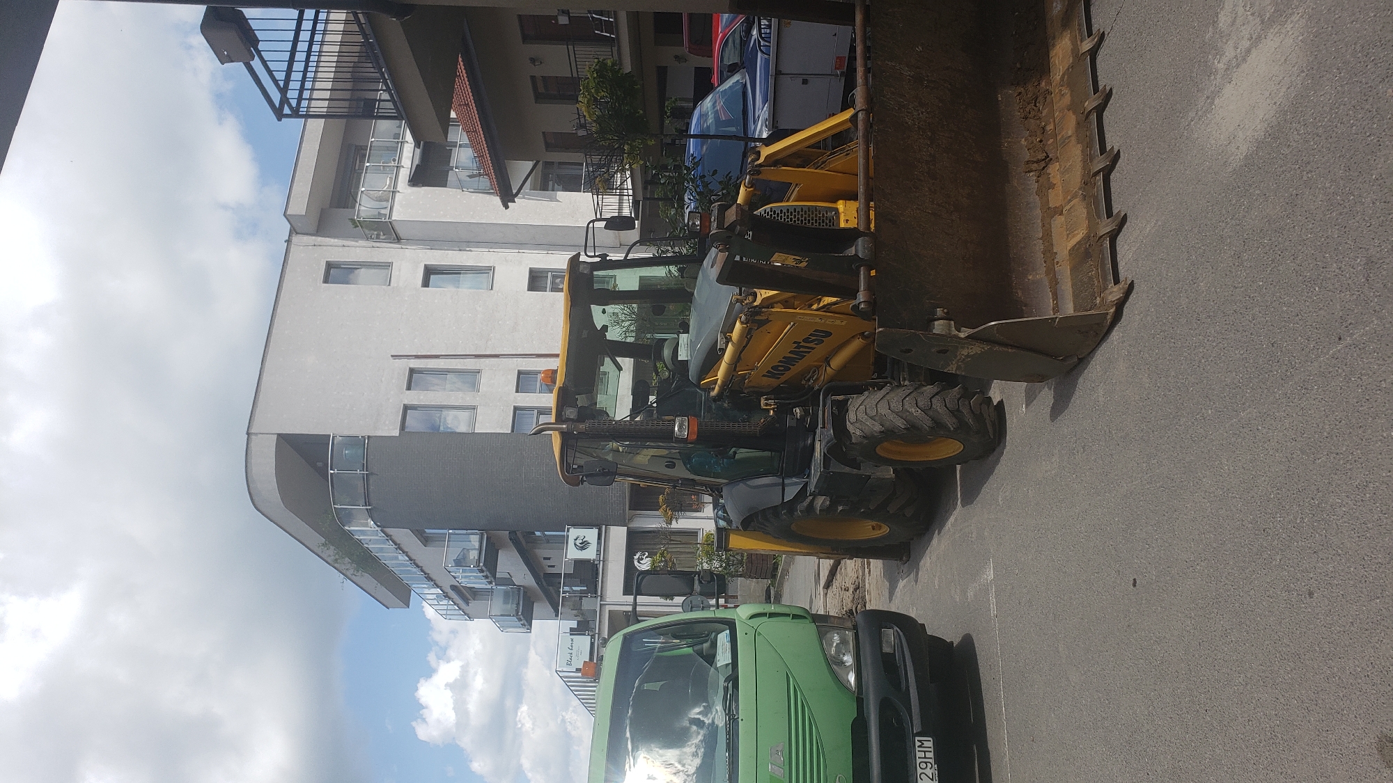 The image shows a yellow Komatsu backhoe loader parked on asphalt pavement in front of a modern building with balconies. A green truck is parked to the left of the backhoe. The building appears to be an apartment complex or hotel. The sky is cloudy. The scene is consistent with an urban environment, likely a street or parking lot in Prešov, Slovakia. There are no people visible in the image. The backhoe has its front loader lowered to the ground, and its rear digging arm is also in a lowered position. The truck has its headlights on and the license plate is partially visible. The building has a distinctive curved upper section.