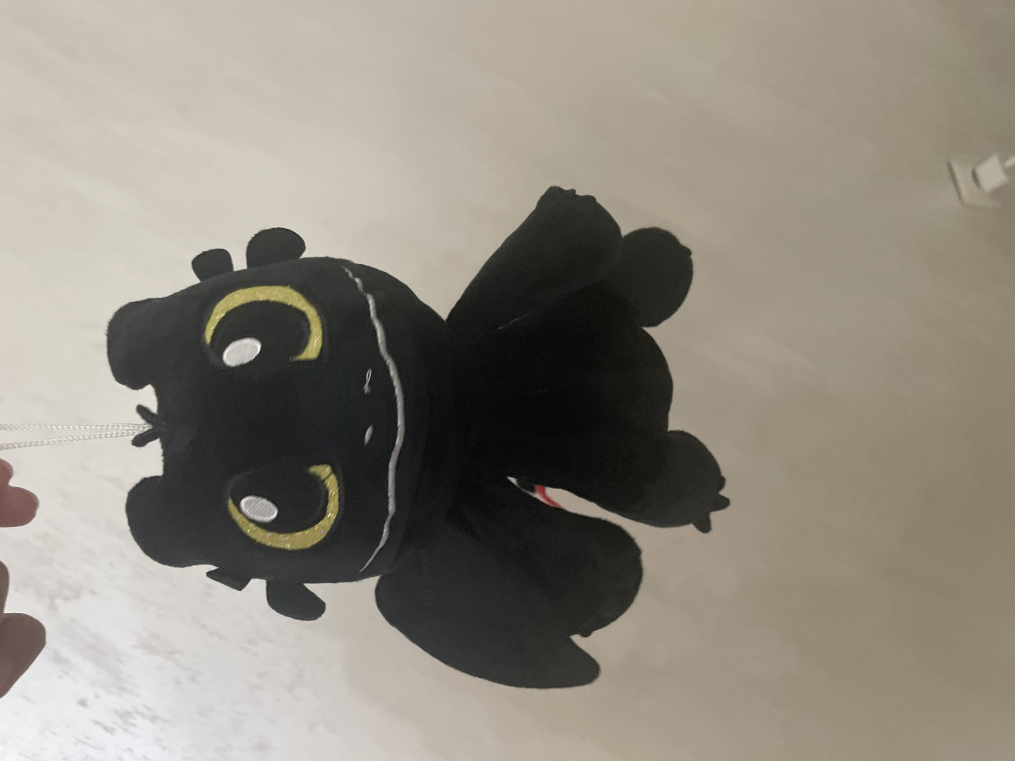 The scene is set indoors, in the city of Ihtiman, Bulgaria, and features a black plush toy, resembling the character Toothless from "How to Train Your Dragon," suspended against a light-toned wall. The plush toy exhibits prominent yellow glitter-textured irises with white pupils, a thin white embroidered smile, small dorsal wings, and minor horn-like protrusions on its head. It is suspended by a slender white string or ribbon, held by two human fingers visible at the top center. The background wall is plain and light-colored, displaying a subtle textural variation. In the lower right quadrant, a white electrical wall outlet is visible, containing a white plug. Illumination suggests a well-lit indoor environment.