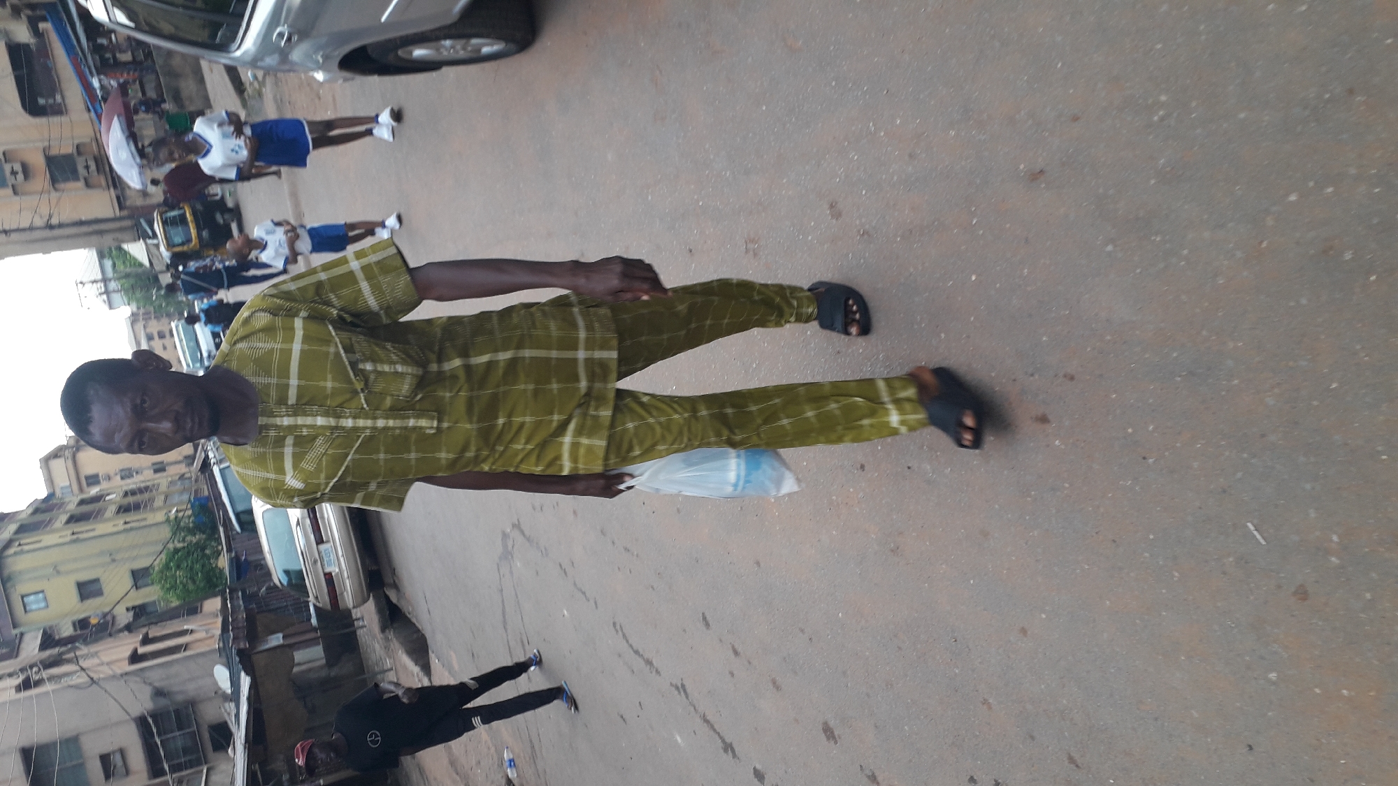 The image depicts a man standing on a street in what appears to be Onitsha, Nigeria, judging by the architectural style and infrastructure visible. He is wearing a green, checkered, traditional African outfit and dark-colored sandals. He is holding a plastic bag. In the background, there are school children in uniform, pedestrians, buildings, cars, and what looks like a tricycle (keke). The street surface is paved and appears worn. There's another person in the lower-left corner wearing all-black clothing. The setting appears to be a typical urban street scene in Onitsha.