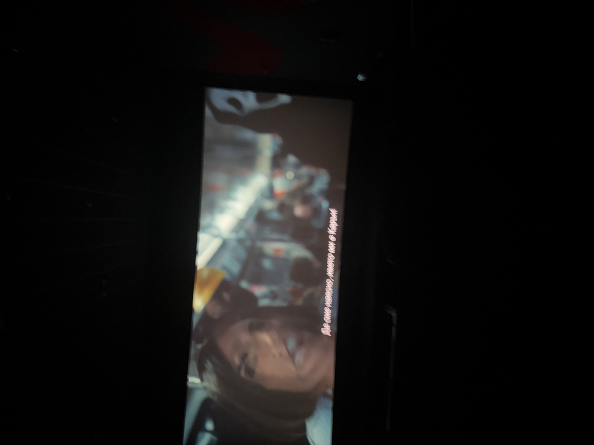 The image displays a dimly lit cinema screen showing a scene from a movie. The screen is framed by black borders and the surrounding environment is dark, suggesting the lights are off for the movie. On the screen, the faces of two individuals are visible. The person on the left, a woman with dark hair, appears to be speaking, with her mouth open. Behind her, another person wearing a yellow hard hat is partially visible. To the right, a silhouette of the iconic Ghostface mask from the "Scream" franchise is discernible, along with other blurry figures in the background. White text in a foreign language, likely Bulgarian given the request for a location in Ruse, Bulgaria, is present at the bottom of the screen. The image does not contain any information that could pinpoint its location to the city of Ruse.