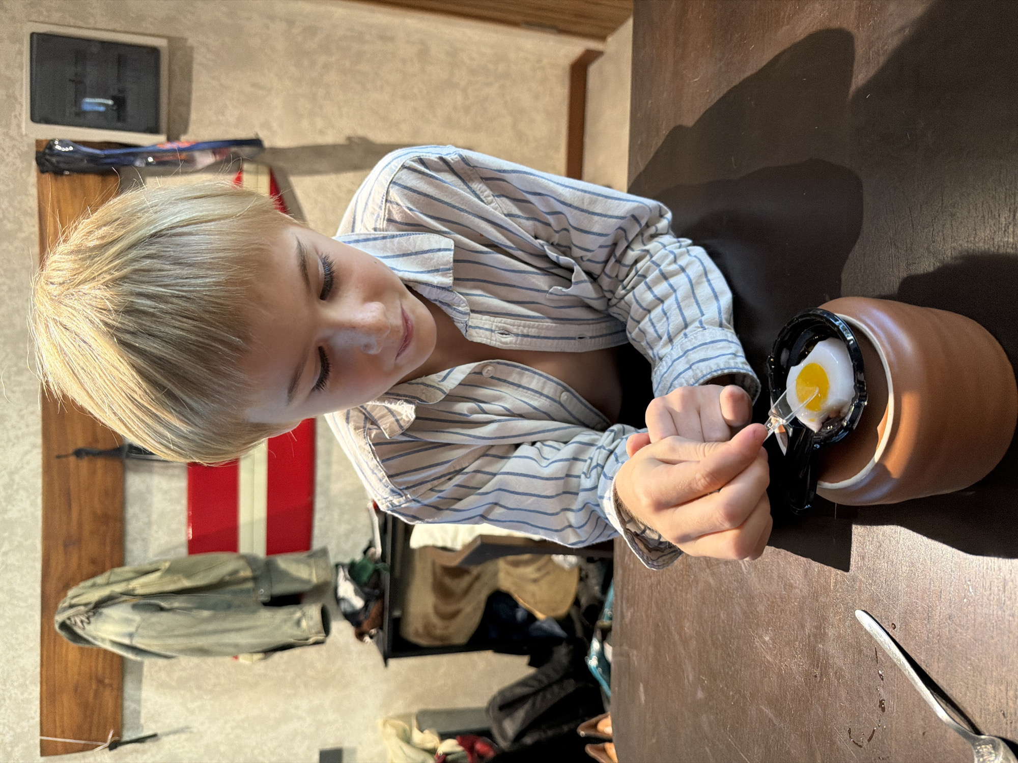 A young fair-skinned person with short blonde hair, wearing a light blue and white striped shirt, sits at a dark brown wooden table. The person is looking down at a small, brown, cup-like container with a black rim and handle that holds a single egg. A yellow yolk is visible on top of the egg. The person holds a small, clear plastic spoon in their right hand, poised to eat the egg. To the right of the cup is a silver spoon on the table. The background includes a wall with a painting or decorative item of red and white stripes. There's also clothing on a rack. This scene is likely taking place in an indoor setting, such as a home or restaurant. While the image may be taken in Kuta Selatan, Indonesia, there is insufficient evidence to confirm this location.