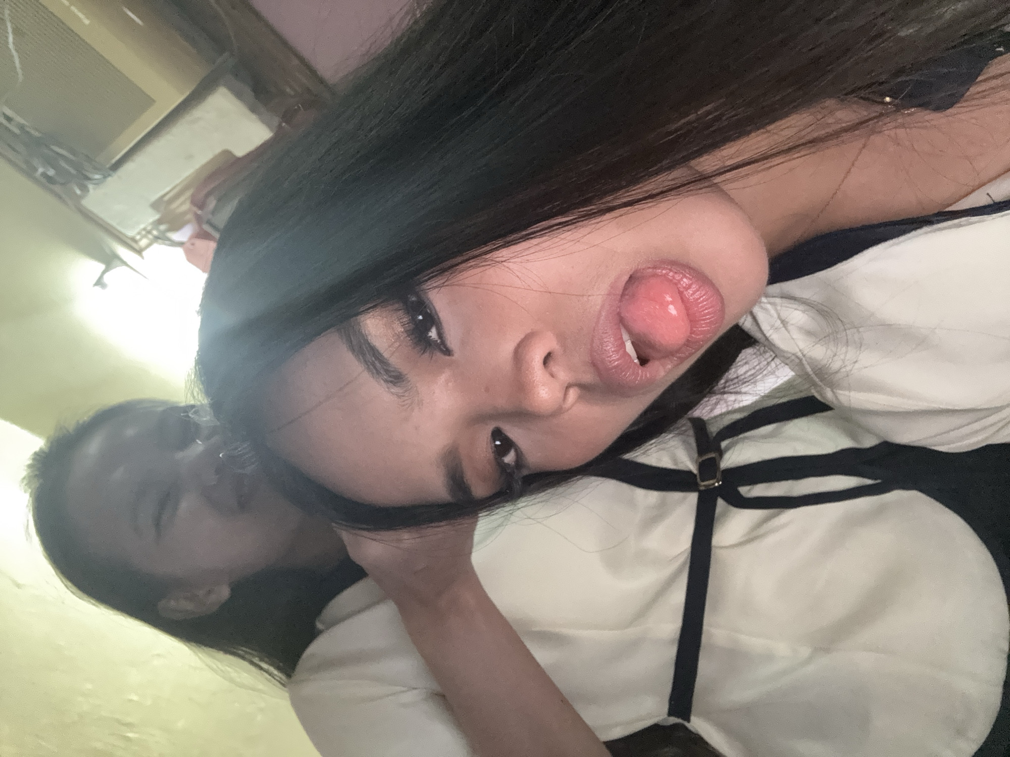 The image shows two young adult women in a close-up selfie. The woman in the foreground is angled towards the camera, sticking her tongue out with her mouth open. She has long, dark hair, heavy eye makeup, and is wearing a white top with black trim and a buckle. Behind her, another young woman with dark hair is visible, with her face contorted in a playful or teasing expression. Her left arm is extended, with her hand resting on the other woman's head. The background is dimly lit and appears to be an indoor setting, possibly a home, with a wall painted a pale yellow and some indistinct objects on a shelf in the upper right corner. There is no specific identifiable landmark or characteristic that would pinpoint a location within Davao City, Philippines. The overall scene is informal and suggestive of a lighthearted moment between friends.