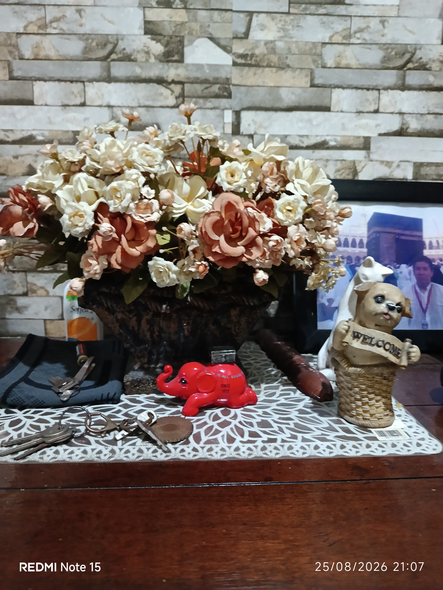 This is a close-up, eye-level, medium shot of a wooden table with various objects on it, set against a wall with stacked stone-like tiling. The primary subjects are a floral arrangement in a woven basket, a small red elephant figurine, a pair of keys, and a decorative dog figurine holding a "WELCOME" banner.
The floral arrangement consists of numerous artificial flowers in shades of cream, beige, and muted rose. The flowers are densely packed in a dark brown, textured basket. To the left of the basket, partially obscured, is a small white bottle with orange labeling, possibly a cleaning product.
In front of the floral arrangement, a bright red, molded elephant figurine is positioned. It appears to be made of plastic and has its trunk raised. To the left of the elephant, a set of keys, including a few house keys and a single car key, are laid out on a white, lace-like placemat. The keys are made of metal and appear aged. A pair of small scissors is also visible near the keys, resting on a dark gray fabric item that might be a shoe or a pouch.
To the right of the elephant, a dog figurine made of ceramic or resin is prominently displayed. The dog has light brown fur and is wearing a blue ribbon around its neck. It is holding a tan banner with the word "WELCOME" written on it in dark lettering. Behind the dog, a white cat figurine can be seen.
Further to the right, a framed photograph is partially visible, showing the Kaaba in Mecca, Saudi Arabia, with a crowd of people in the background.
The table surface is made of dark, polished wood. The overall lighting suggests an indoor setting, possibly a home or a shop. The image is dated "25/08/2026 21:07" and is identified as "REDMI Note 15" in the lower left corner, indicating it was taken with a Redmi Note 15 mobile phone. There is no specific information about the location in Keboananom, Indonesia, within the image itself.