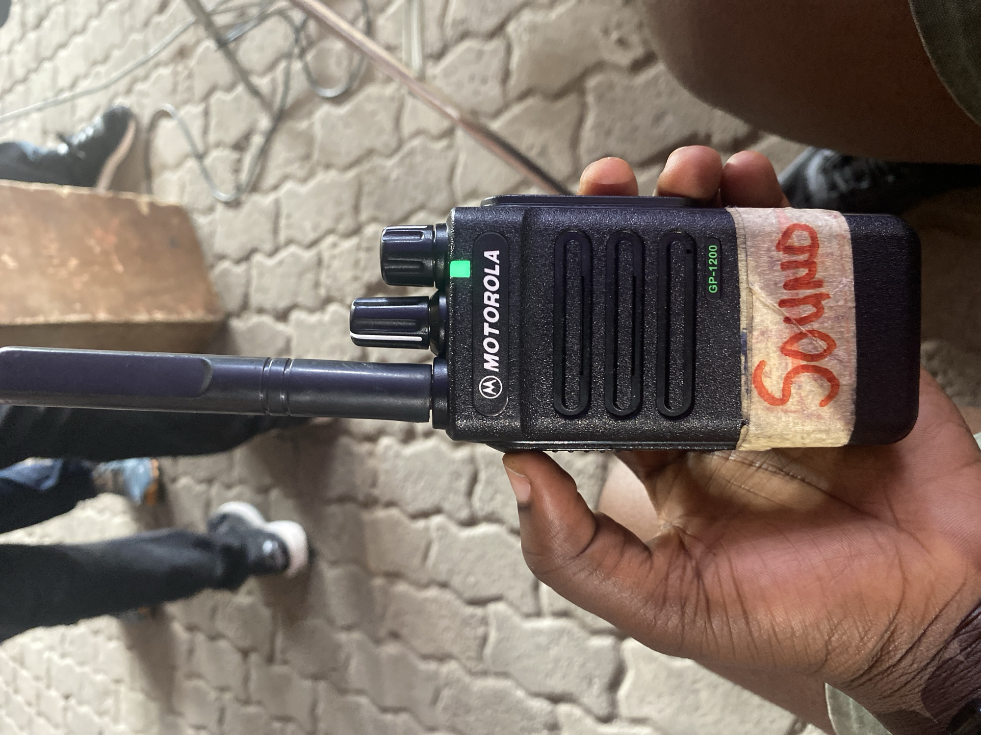 A person is holding a Motorola GP-1200 walkie-talkie with the word "Sound" written in red on tape. The background shows pavement stones and two sets of legs and feet wearing sneakers. The location is Lagos, Nigeria.