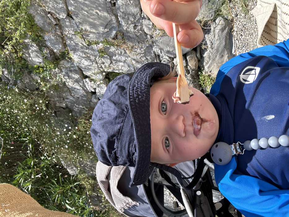 A young child, estimated to be a toddler, with fair skin and blue eyes, is centered in the frame, looking forward. The child wears a navy blue bucket hat and a blue long-sleeved rash guard featuring a white logo on the chest. A grey beaded pacifier clip is attached to the rash guard. Dark brown ice cream or chocolate is smeared around the child's mouth and drips onto the chin and rash guard. From the right, an adult's hand offers a small wooden spoon containing a scoop of light-colored ice cream topped with a dark brown substance. To the left, a portion of a grey stroller with a beige blanket draped over it is visible. The foreground consists of rocky and pebbly ground. The background is dominated by a stone wall covered with green foliage and small white flowers. A part of a straw hat is visible in the upper left corner. This outdoor scene is located in Abbadia Lariana, Italy.