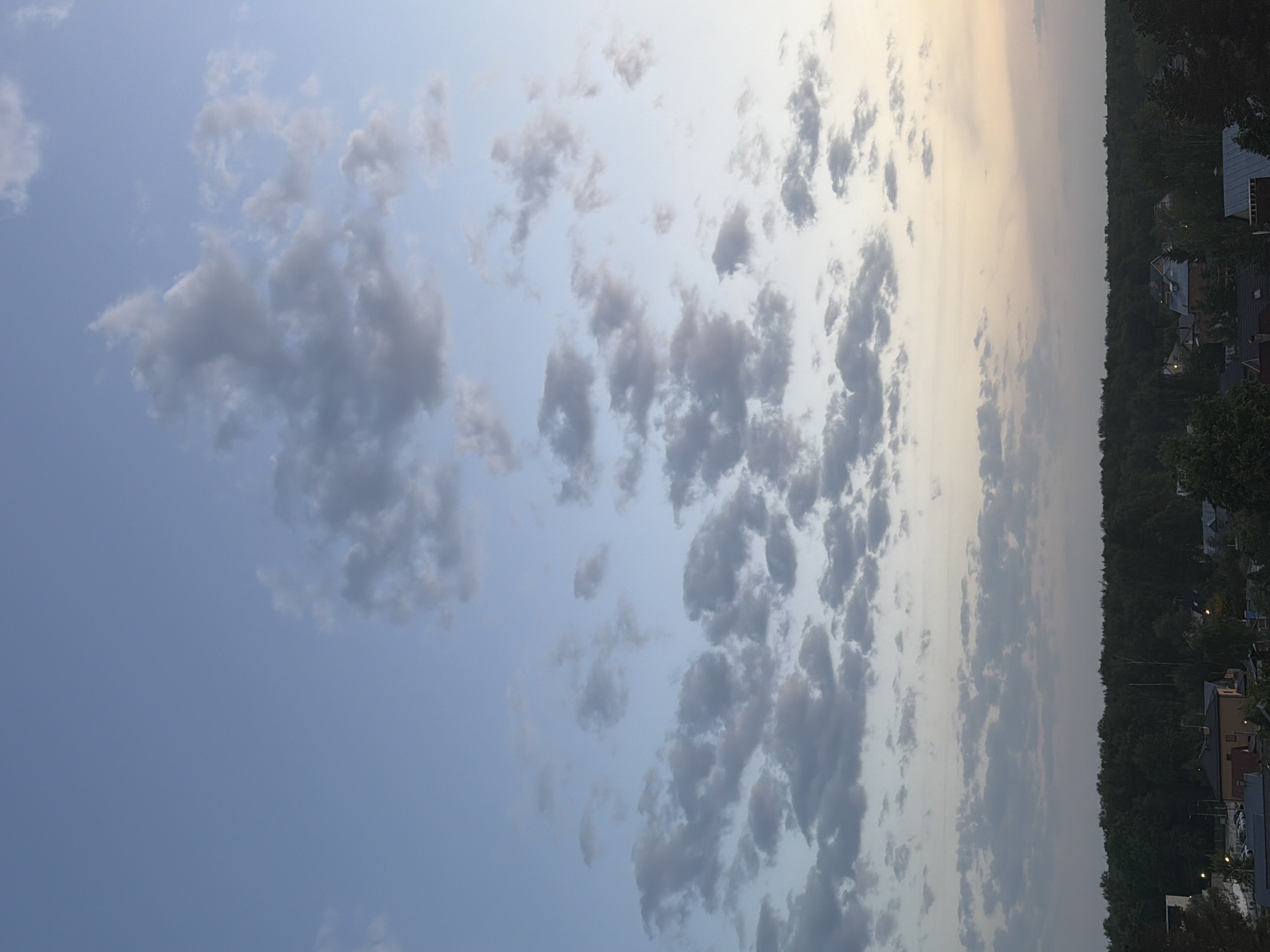 The image shows a sky filled with altocumulus clouds. The sky transitions from a lighter blue on the left to a more muted tone on the right, near the horizon. At the far right, there is a line of trees and buildings suggesting the edge of a city or town. The location is Unknown, Unknown.