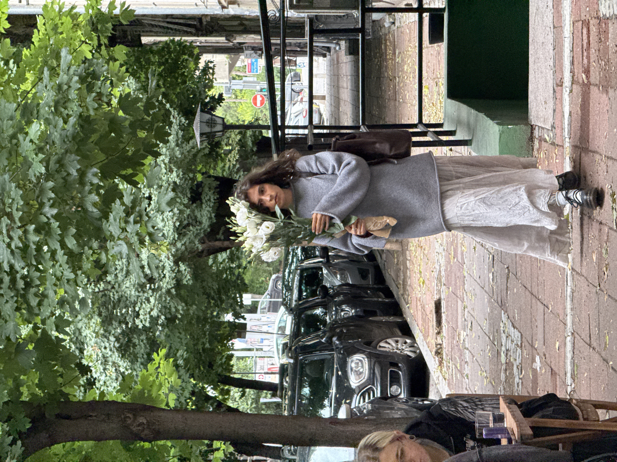 A young woman walks down a cobblestone sidewalk in Belgrade, Serbia, carrying a large bouquet of white flowers. She is wearing a grey oversized sweater and a long, flowing white skirt. Her hair is dark and wavy, and she has a serious expression on her face. A brown backpack is slung over her right shoulder. The sidewalk is lined with parked cars, and trees with green leaves overhang the street. In the background, a street lamp is visible, as well as a pedestrian crossing with traffic signals. To the left of the woman, a person sits at a small wooden table outside a cafe, with a glass of water and a purple object on the table. The overall scene suggests a quiet moment in a European city.