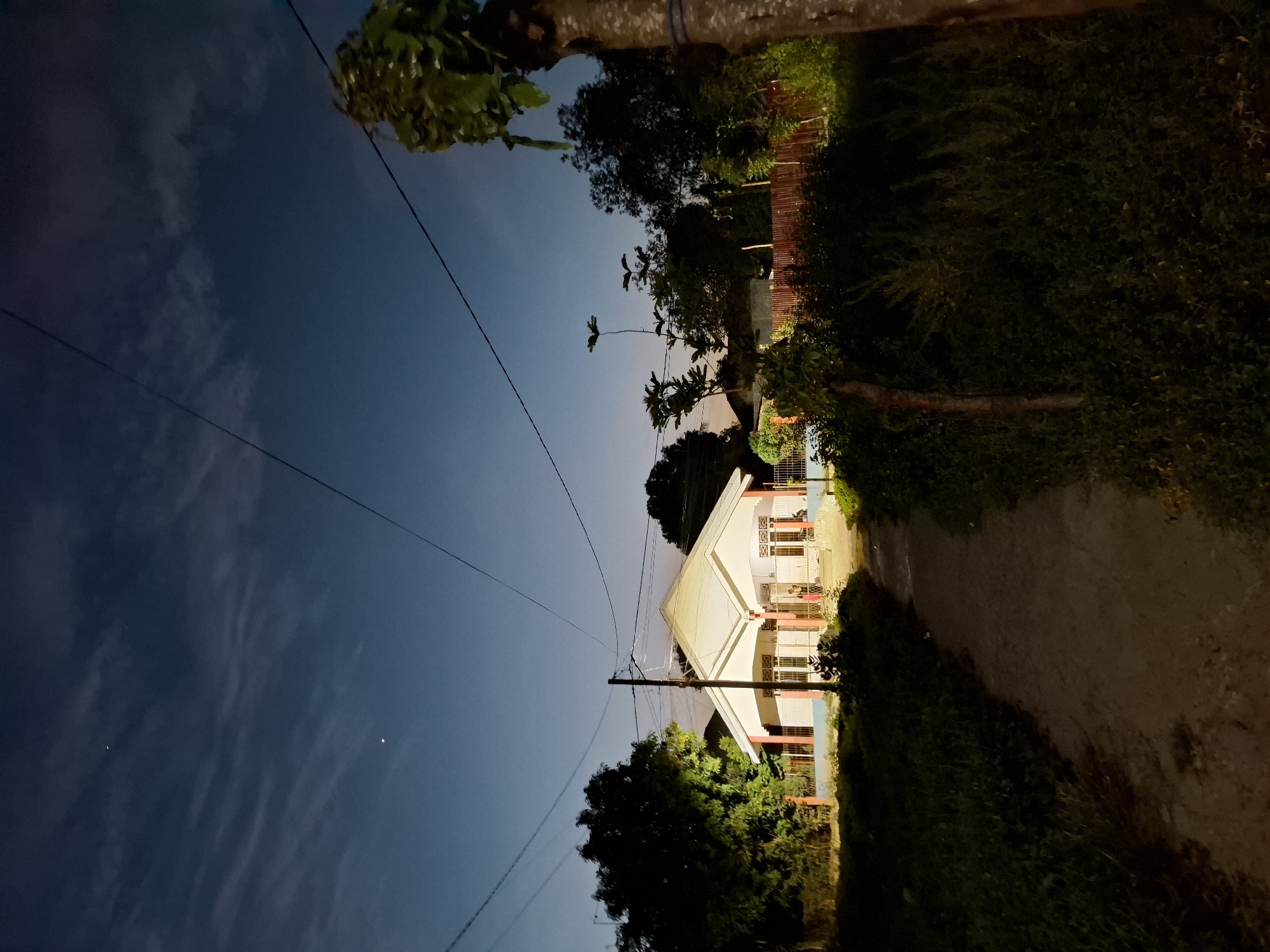 A slightly tilted, long shot shows a two-story house at night, possibly in Lhokseumawe, Indonesia, based on the request. The house has a white exterior with red columns and appears to be lit from within. The sky above is dark with some visible cloud cover. Power lines stretch across the upper portion of the image. Vegetation, including trees and bushes, frames the sides and bottom of the house. A dirt path runs along the right side of the house. The overall scene has a dimly lit, nighttime atmosphere.