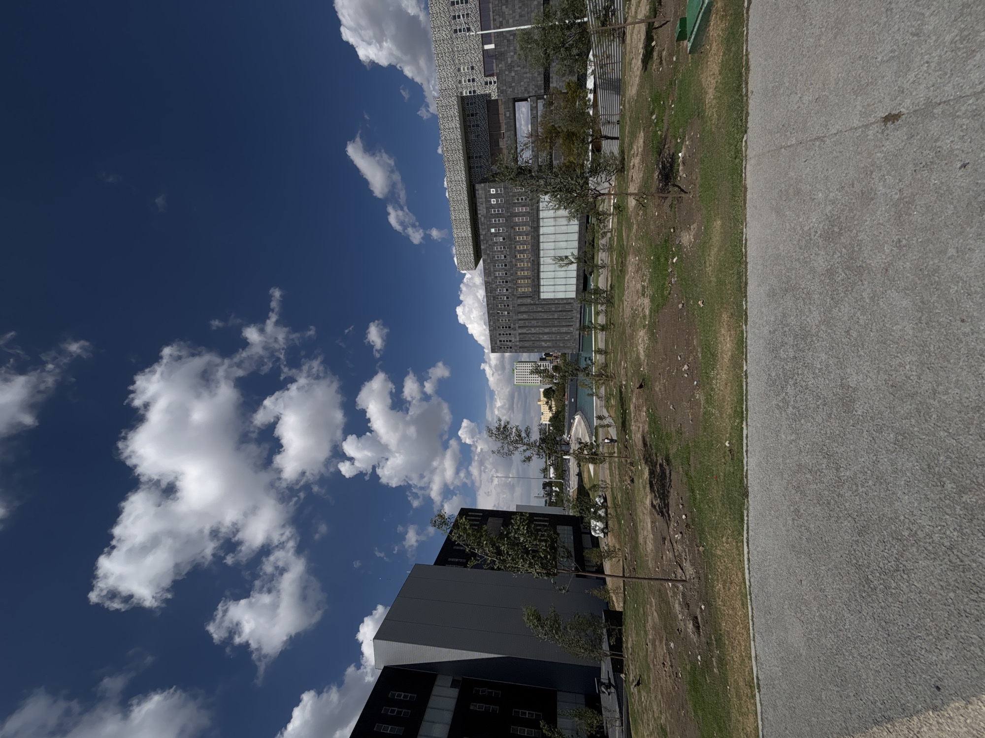 The image depicts a low-angle view of a cityscape on a sunny day with a blue sky and scattered white clouds. The perspective is rotated ninety degrees clockwise. Multiple tall, modern buildings with glass and concrete facades dominate the horizon, with some appearing to be residential and others commercial. A grassy area with trees and sparse vegetation extends from the bottom of the frame towards the buildings. A wide concrete walkway or sidewalk runs along the right side of the image.
Based on the architectural style and visual cues, this scene likely represents a contemporary urban development in Puebla, Mexico. There are no people visible in the image.