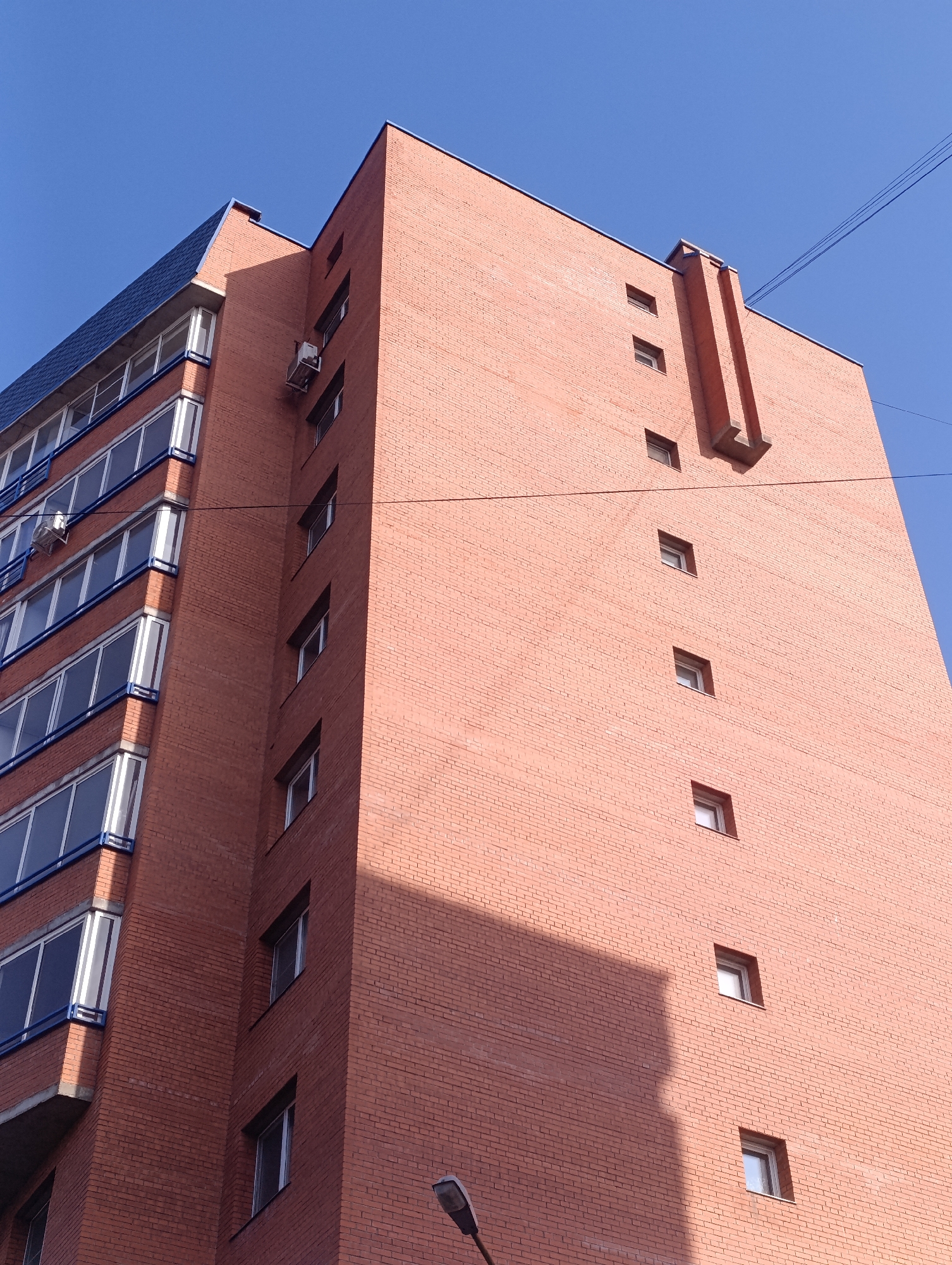 The image shows a tall, red brick building under a clear blue sky. The building appears to be located in Krasnoyarsk, Russia, based on architectural style. The left side of the building features multiple stories with balconies and blue accents. The right side is a solid brick wall with sporadically placed small windows and a vertical, decorative architectural element near the top. A streetlight is visible at the bottom of the frame, and overhead wires cross the upper portion. A shadow is cast on the lower portion of the brick wall. No people are visible.