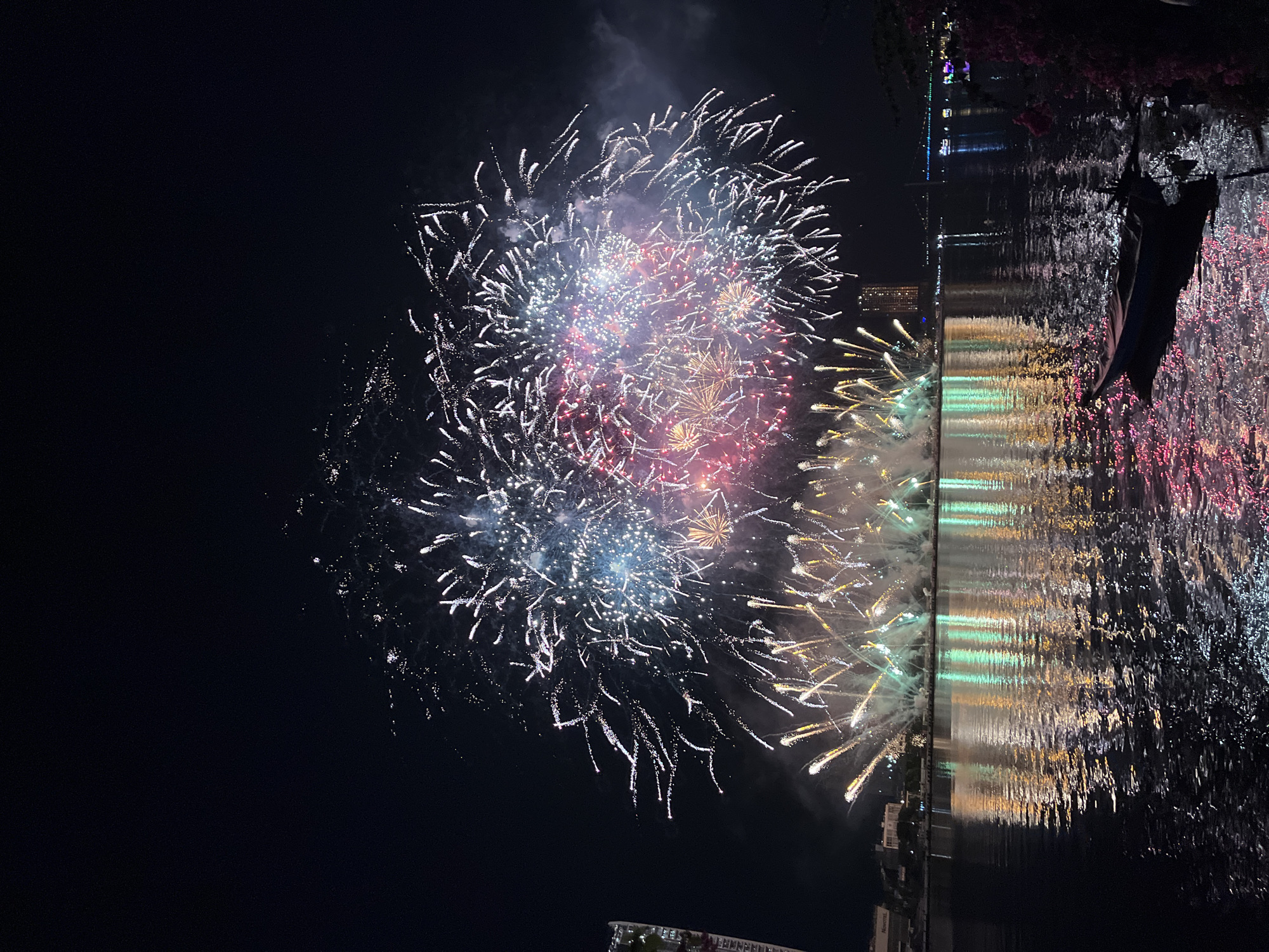 A nighttime photograph captures a large-scale fireworks display illuminating the sky above the Han River in Đà Nẵng, Vietnam. The upper two-thirds of the image are dominated by numerous fireworks exploding in white, blue, red, and gold hues, with visible smoke plumes. Lower fireworks burst from the water's edge, emitting initial upward streaks of golden and green light. The lower half of the image features the dark river, its surface displaying rippled reflections of the fireworks in vibrant streaks of green, gold, pink, blue, and white. Along the distant riverbank, multiple illuminated buildings are visible, including a tall high-rise structure on the left and a building clearly marked "NOVOTEL" further right. An illuminated bridge spans the river in the far background. In the immediate bottom-right foreground, a small, dark boat rests on the water, partially obscured by dark foliage exhibiting faint pink highlights from the fireworks. No individuals are discernible within the scene.