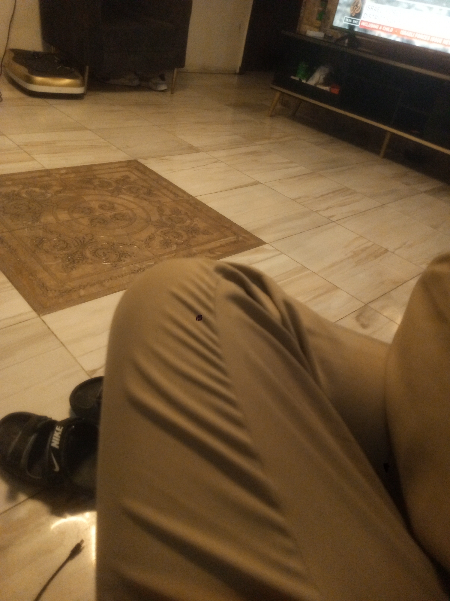 The image depicts a person sitting indoors. They are wearing tan-colored pants and black Nike sandals. A rug with an ornate pattern is partially visible on the tiled floor. In the background, there is a dark-colored cabinet and a television set. The location is Gwarinpa, Nigeria.