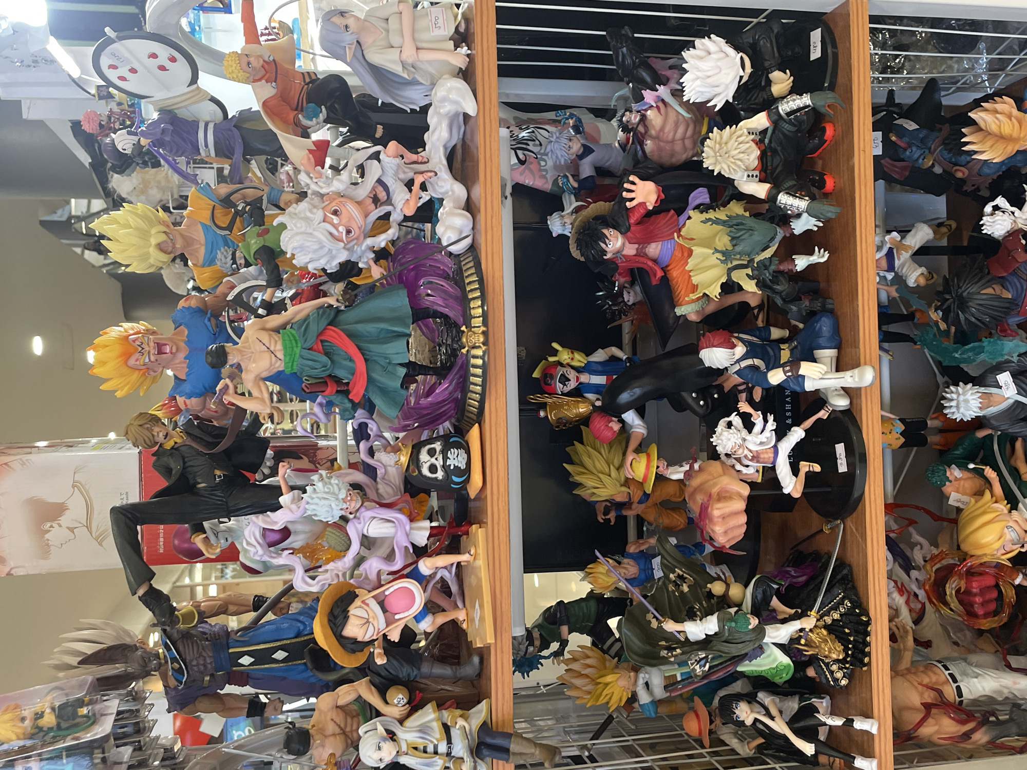 The image shows a shelf of anime figurines, likely in a store, with numerous characters from popular series like Dragon Ball Z, One Piece, and others. The figurines are arranged on multiple shelves, with some figures larger than others. The top shelf features characters such as Beerus, Frieza, Goku, Vegeta, and Sanji. The middle shelf displays characters like Monkey D. Luffy, Roronoa Zoro, and several other anime characters. The bottom shelf has more figurines, including some from Naruto and My Hero Academia. The overall environment suggests a retail setting, possibly a toy or collectible shop. There are no people visible in the image, only the static displays of figurines. The lighting appears to be artificial, typical of indoor retail environments. The figurines are made of plastic and are highly detailed, representing various characters in dynamic poses. There is no specific information within the image that would indicate its location in Davao City, Philippines.