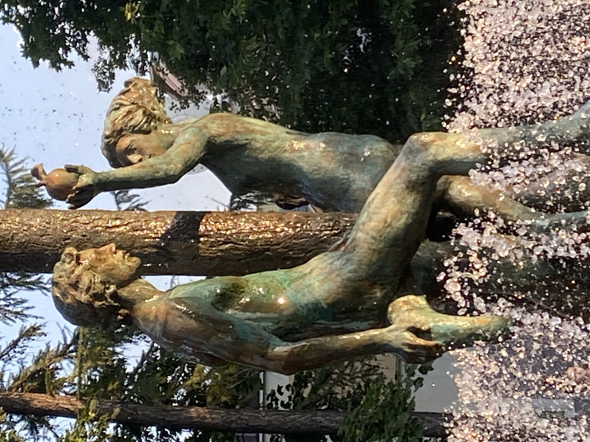 The image shows a bronze sculpture of a nude couple by a fountain. The male figure is on the left, leaning against a tree trunk, and the female figure is on the right, holding an apple aloft. Both figures appear to be made of bronze and are wet from the fountain spray. The sculpture is located in Gorky Park, Moscow, Russia. The background consists of trees and a building. The fountain water is creating a shimmering effect around the base of the sculpture. The lighting suggests it is daytime, possibly late afternoon given the warm light.