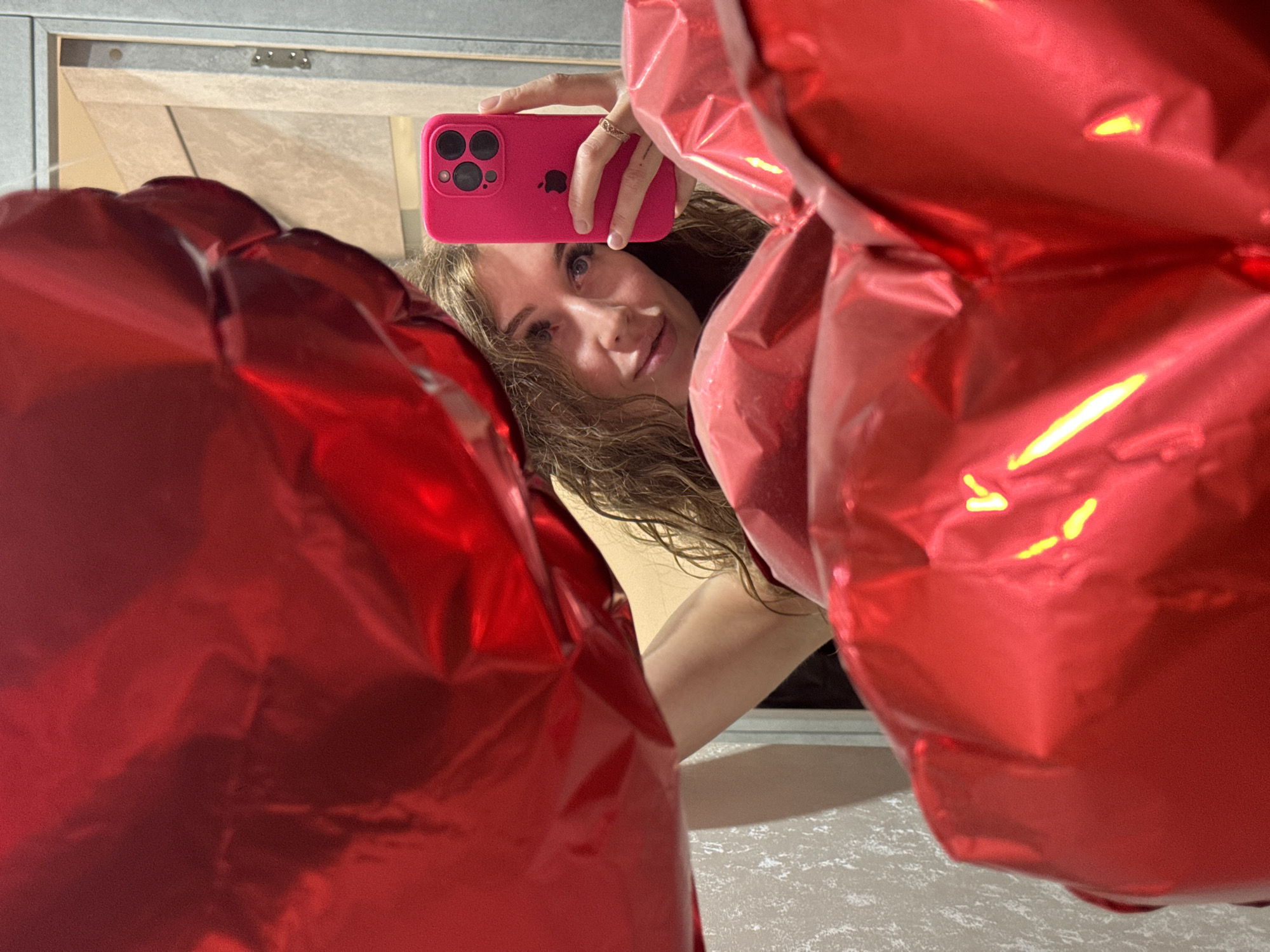 A woman is taking a selfie in what appears to be a bathroom in Chaikovsky, Russia. The woman is holding a pink smartphone in her right hand and is positioned behind two large, red, heart-shaped balloons. Her face is visible between the balloons, and she is looking directly at the camera. The background includes a mirror, a white countertop, and possibly a cabinet above the mirror.