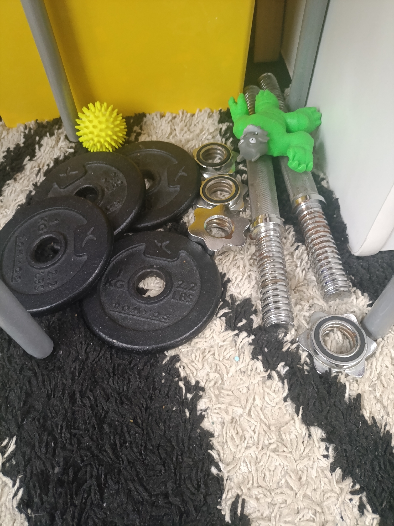 The image shows a collection of gym equipment scattered on a black and white shag rug. The items include four black weight plates, two dumbbell bars with securing collars, and a yellow spiky ball. There is a green action figure resting on the dumbbell bars. The background includes a yellow storage bin and a white object, possibly a piece of furniture. The location is presumed to be indoors. There is no information to confirm that this photo was taken in Krasnodar, Russia.
