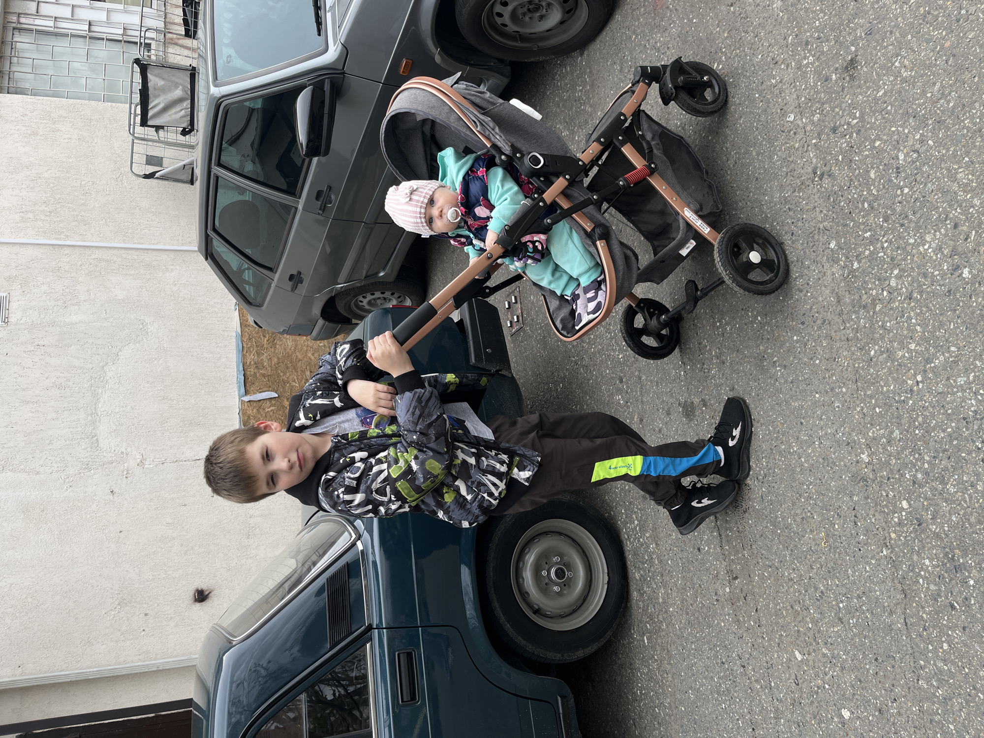 A young boy in a patterned jacket leans against a dark green car, while next to him, a baby sits in a stroller. The setting appears to be a street, based on the asphalt and the presence of other cars, one of which is gray. The location is Sochi, Russia. The image seems to depict a common family scene in an urban environment.