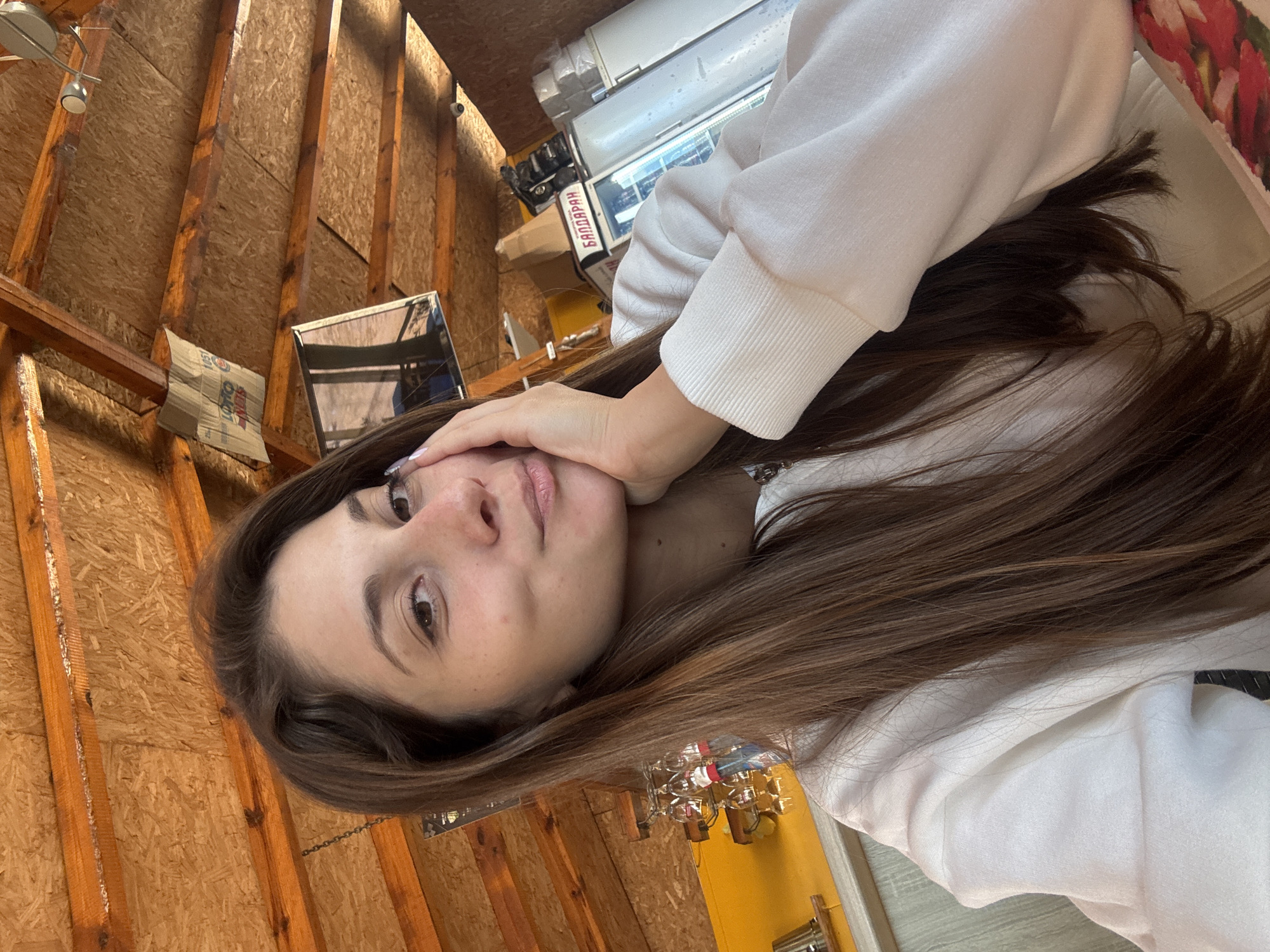 A woman with long, brown hair rests her face on her left hand while standing indoors. She wears a light-colored sweatshirt. The background features unfinished wood walls and ceiling, as well as various objects including lights, boxes, and a display case with Bulgarian writing ("БАЛДАРАН"). The location is likely a shop or storage area in Sozopol, Bulgaria, given the Bulgarian text and the rustic construction style.