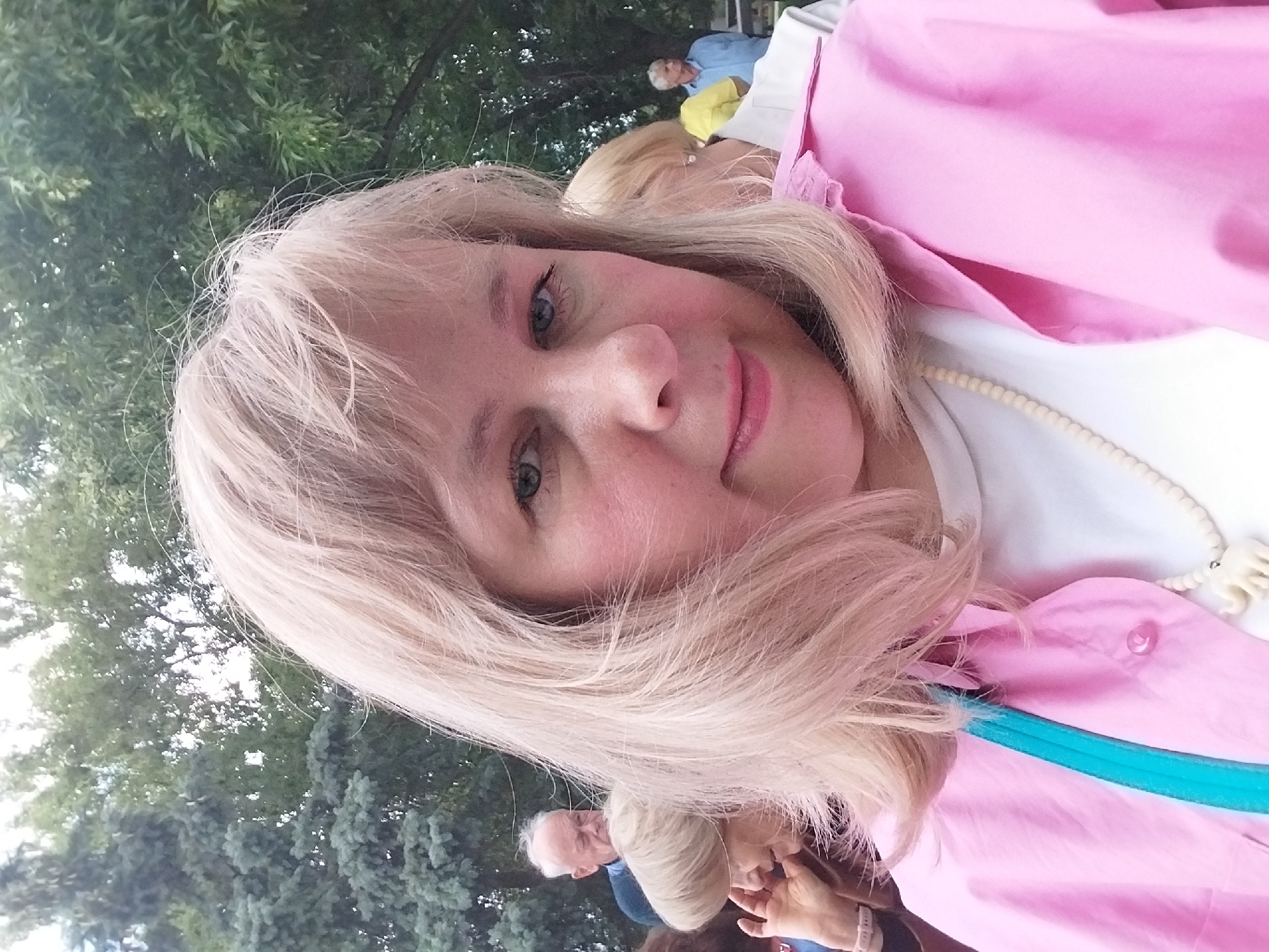 A blonde woman with blue eyes and fair skin smiles gently in a close-up, selfie-style portrait. Her shoulder-length hair is light blonde, and she has rosy cheeks. She is wearing a bright pink jacket or shirt over a white top, and a long, cream-colored beaded necklace with larger, stylized elements resembling small bones or teeth.
The setting appears to be outdoors, likely a park or public gathering, with lush green trees filling the soft-focus background. Several other people are partially visible behind her, including individuals wearing yellow and blue clothing, and another person with white hair. The lighting is soft and diffused, suggesting it is daytime, possibly overcast or in a shaded area. The woman’s expression conveys a pleasant and relaxed mood. No text is visible in the image. This scene takes place in Orenburg, Russia.