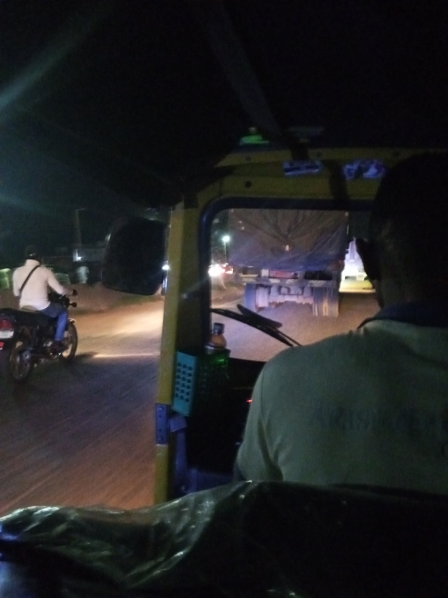 The image depicts a scene from the interior of a yellow auto-rickshaw at night, likely in Lokoja, Nigeria. In the foreground, the back of the auto-rickshaw driver, wearing a light-colored shirt with some text on the back, is visible. The driver is facing forward, looking towards the road ahead. A dark, possibly plastic-wrapped object is on the floor of the auto-rickshaw.
Through the windshield, a motorcycle with a rider is seen moving away from the camera. The rider is wearing a white shirt and blue jeans, with a bag slung over their shoulder. The motorcycle's headlight is on, illuminating the road. The road surface appears to be asphalt, with some unevenness and possibly dust.
In the background, a large truck covered with a blue tarp is parked or moving slowly. Other vehicles with illuminated headlights and taillights are visible in the distance, suggesting a busy road. Streetlights can be seen in the far background, casting a yellowish glow. The overall environment is dark, characteristic of nighttime in an urban setting. The image captures a fleeting moment of travel, emphasizing the darkness and the artificial lights of the city.