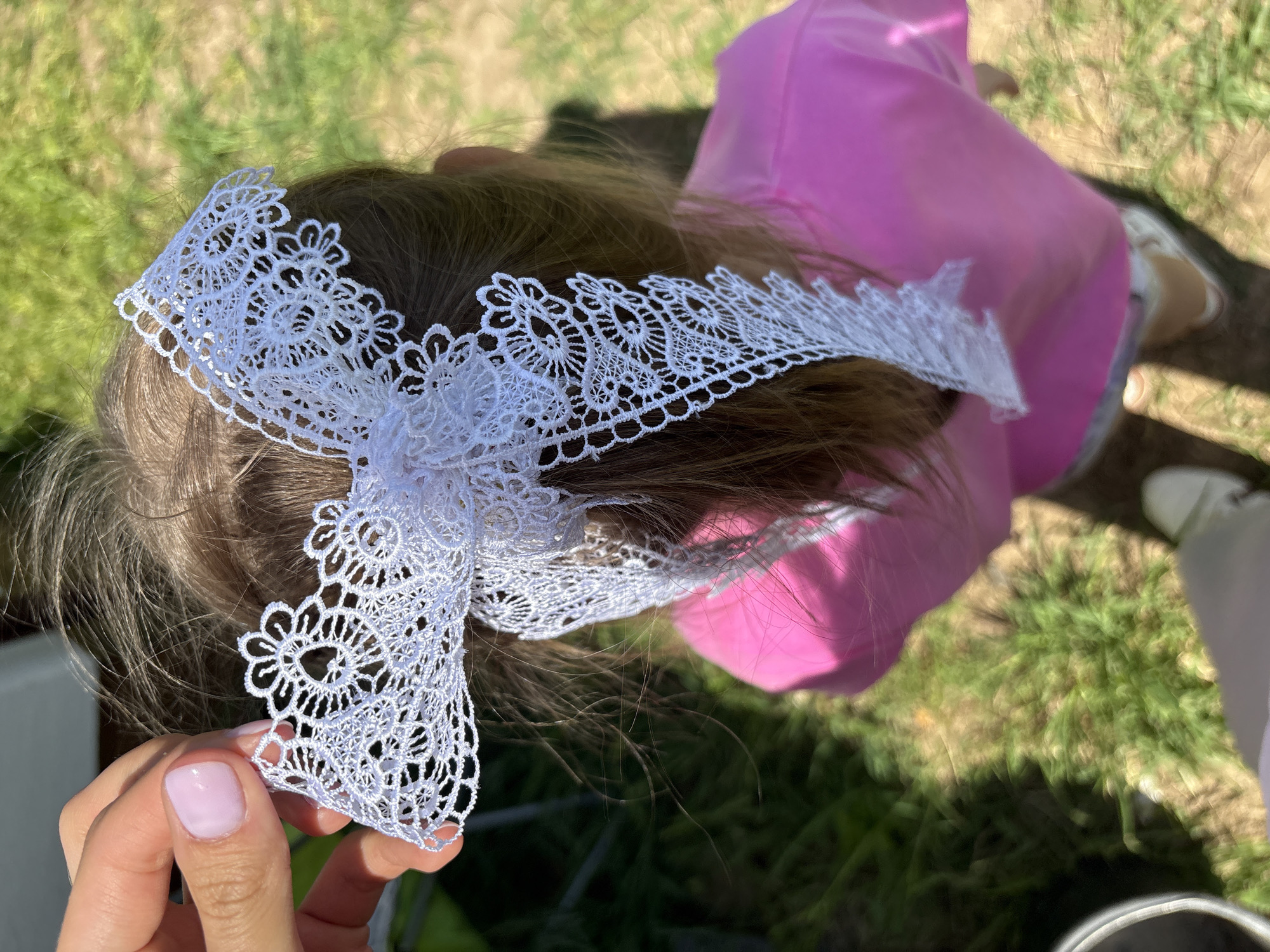 A high-angle shot depicts a child wearing a white lace headband. The headband, held by a person's hand with manicured nails, features a floral pattern. The child, positioned outdoors in a grassy area, is wearing a pink garment. The location appears to be Sibir', Russia, given the prompt.