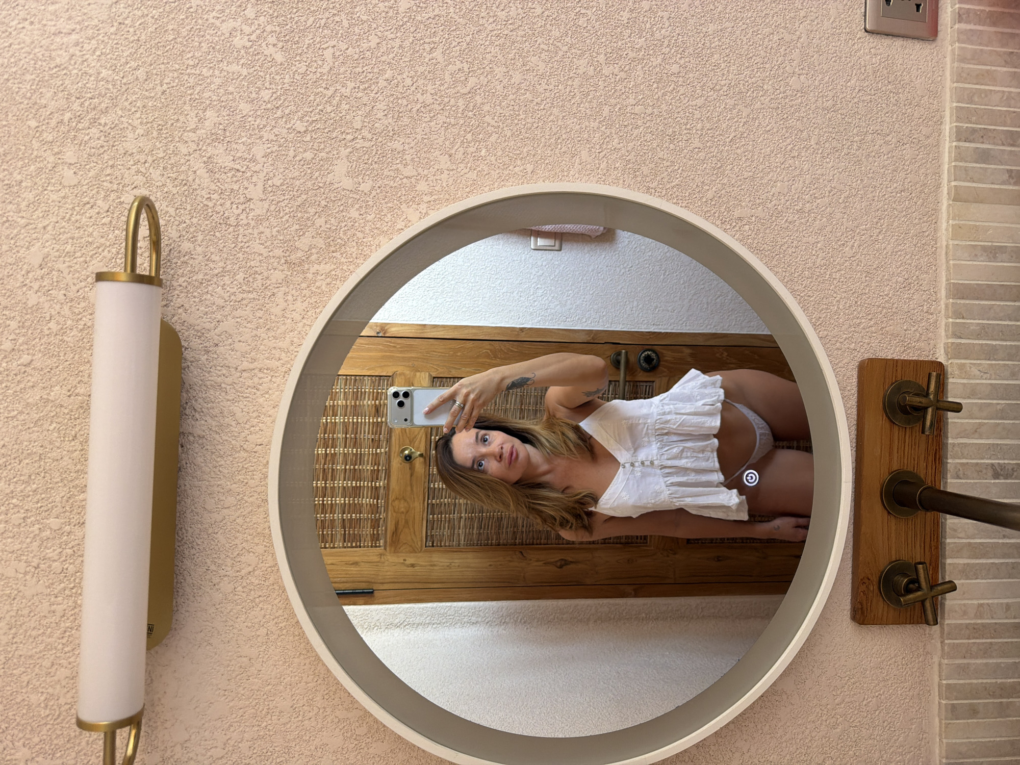 A young woman is captured taking a mirror selfie in what appears to be a bathroom or vanity area, likely in Mengwi, Indonesia. The image is taken from the perspective of the woman holding a light-colored smartphone, which is reflected in a round, light-framed mirror.
The woman has medium-length brown hair and is looking directly into the camera with a neutral expression. She wears a white, sleeveless top with a ruffled hem and small buttons down the front; white underwear is visible beneath the top. A ring is seen on her left hand, which holds the phone, and she has a small bird-like tattoo on her left inner forearm.
The wall behind the camera has a textured, light peach or beige finish. To the left of the mirror, a wall-mounted light fixture features a brass-colored bracket holding a cylindrical white diffuser; part of the word "NI" is visible on its side. On the right, vintage-style cross-handle faucets and a spout are mounted on a rustic wooden panel. Adjacent to these, a section of wall is covered with horizontally stacked light-colored tiles. A silver electrical outlet is visible in the upper right corner.
Inside the mirror's reflection, a wooden door or cabinet with a woven rattan pattern is seen behind the woman. Below this, a white textured wall is reflected. A circular, glowing white icon resembling a power or restart symbol is superimposed on the reflection of her body on the mirror's surface, indicating an illuminated mirror feature. The overall aesthetic suggests a blend of modern comfort with natural and rustic design elements.