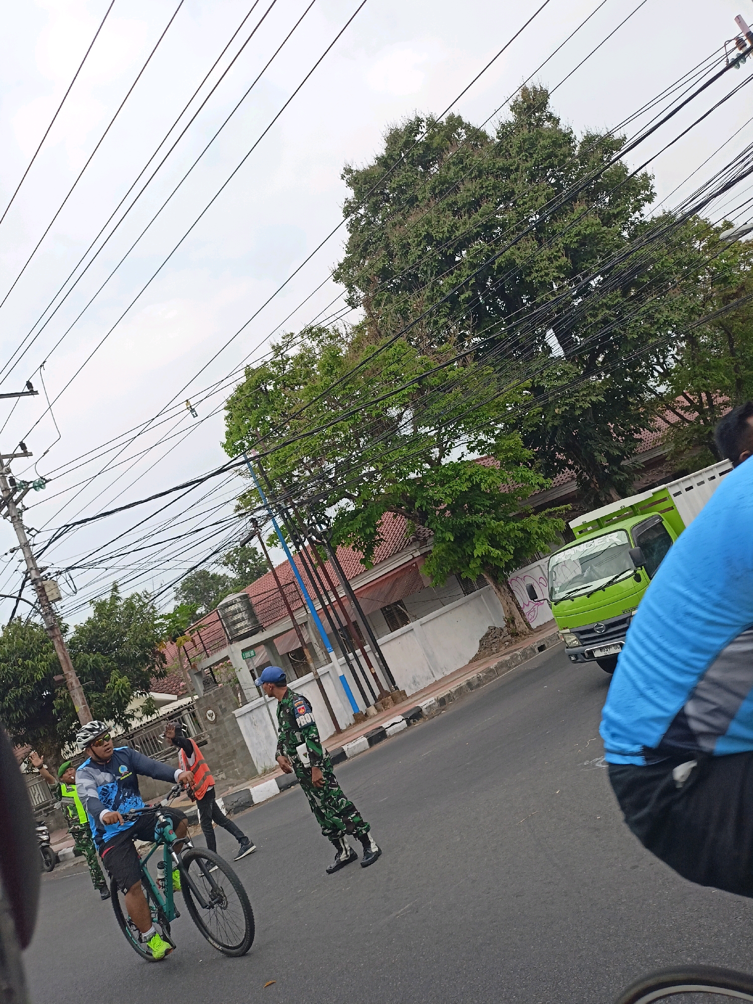 The image depicts a street scene in Magelang, Indonesia, under an overcast sky. A group of people are present, including individuals in military uniform and a person on a bicycle. The person on the bicycle is wearing a helmet, cycling jersey, and shorts. To the right, two individuals in camouflage uniforms appear to be standing or walking. In the background, a small green truck is visible, along with buildings with red tile roofs and a large tree with dense foliage. Utility poles and a complex network of overhead wires are prominent features of the environment. The ground is paved with asphalt. The overall impression is that of a daytime street scene, possibly during a public event or a routine patrol, given the presence of military personnel and cyclists.