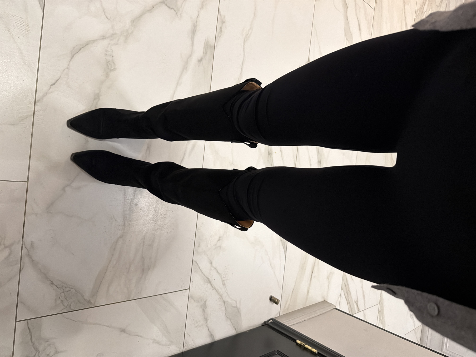 The image presents an overhead, downward-angled view of a person's lower body from the waist down, focusing on their legs and feet. The person is wearing tight-fitting black pants or leggings and knee-high or over-the-calf black boots with pointed toes. A small fragment of a light-colored, possibly grey, garment with a button is visible at the very top right edge, hinting at the wearer's upper attire.
The setting is indoors, featuring a clean, light-colored tiled floor with distinct grey marble-like veining patterns. The tiles are large and arranged with visible grout lines. Towards the bottom right corner, a section of a dark, likely black, door frame or threshold is visible, adorned with metallic hardware, possibly a brass-colored hinge or lock component. A small, metallic cylindrical object, appearing to be a spent bullet casing, lies on the tiled floor near the door frame.
The image captures a static moment, with the person seemingly standing or poised to move. The lighting appears to be artificial, casting no harsh shadows. The overall impression is of an indoor space, possibly an entrance or hallway, within a residence or building in Moscow, Russia. No visible text is present in the image.