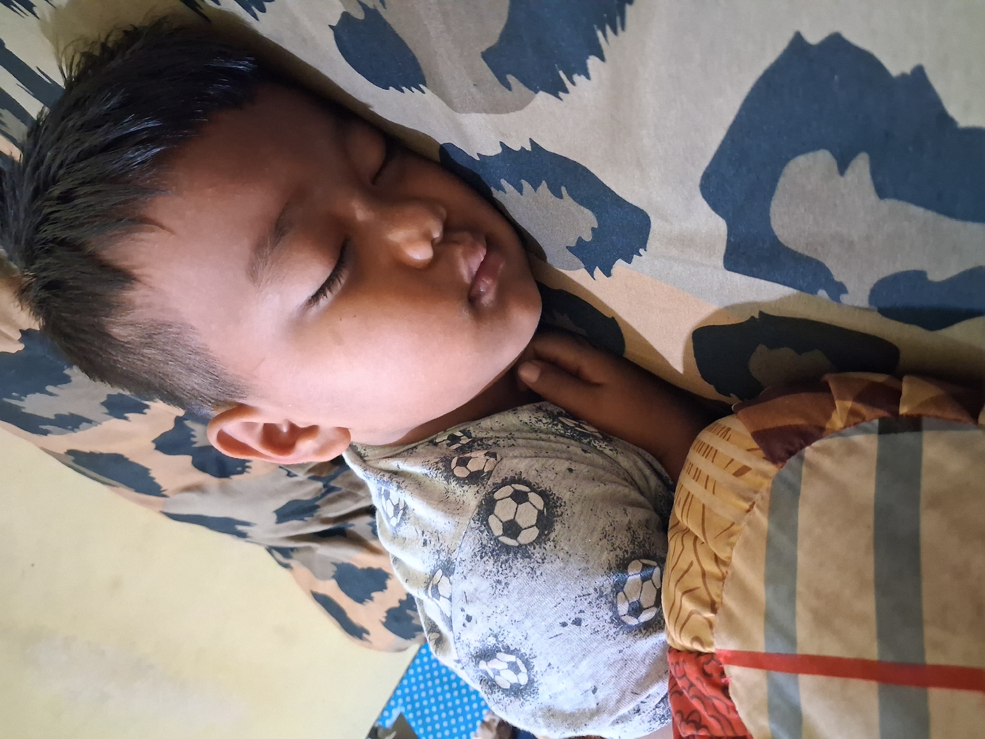 A young child is sleeping soundly on a bed covered with blankets and a pillow. The child is wearing a grey t-shirt with a pattern of soccer balls. The pillow has a camouflage pattern. The child appears to be in a bedroom. This image was taken in Pendawa, Indonesia.