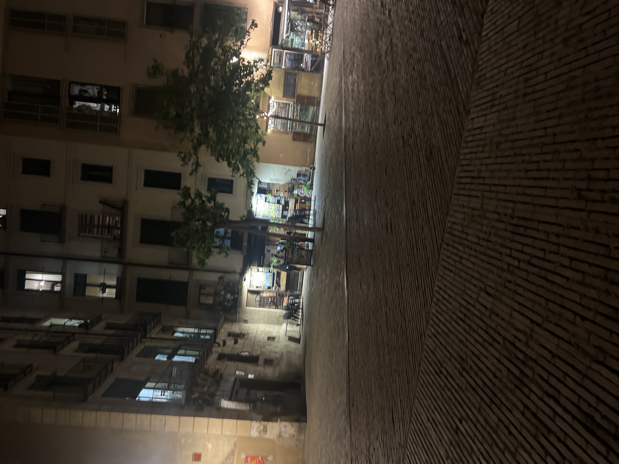 A nighttime photograph captures a narrow urban street or alleyway in a city like Barcelona. The scene is predominantly dark, illuminated by artificial light sources from buildings and a commercial establishment in the background.
On the left side of the street, a tall, multi-story building stands, its facade featuring numerous windows, some of which are brightly lit from within, casting glows and revealing interior reflections. Several windows are equipped with small balcony railings. A leafy tree partially obstructs the view of the building in the mid-ground.
The right side of the street and the ground are paved with a distinctive patterned material, appearing as long, narrow bricks or tiles. These are arranged diagonally, with two sections meeting in a central V-shape pattern.
In the background, at the end of the street, a brightly lit commercial establishment, possibly a bar or restaurant, is visible. It features outdoor seating with tables and chairs, and several blurry figures of people are present, suggesting social activity. Displays or shelves can be discerned through its windows. A smaller tree grows near this establishment.
The image conveys the ambiance of a quiet evening in a historic urban environment. No distinct text is clearly legible from any signs. The architecture and street layout are characteristic of a European city.