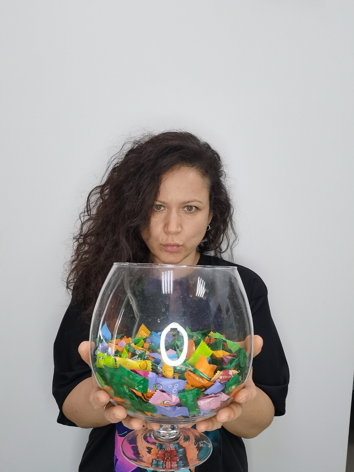 A medium shot shows a woman with long, curly dark hair and fair skin, holding a clear, glass bowl filled with colorful, individually wrapped candies. She is looking directly at the camera with a playful pout. The woman is wearing a black t-shirt with a colorful graphic design on the lower portion, and a silver hoop earring in her left ear. A silver nose ring is visible. The bowl is large and round, with a stemmed base, resembling a fishbowl or a large brandy snifter. The candies are in various colors including green, yellow, orange, purple, and pink, with some wrappers displaying text and graphics that are too small to read clearly. The background is a plain white wall, creating a studio-like setting. The lighting is even, and there are reflections visible on the glass bowl, including a prominent circular reflection. The overall impression is one of casual presentation, possibly for a promotional or personal content. The image was captured in Chaikovsky, Russia.