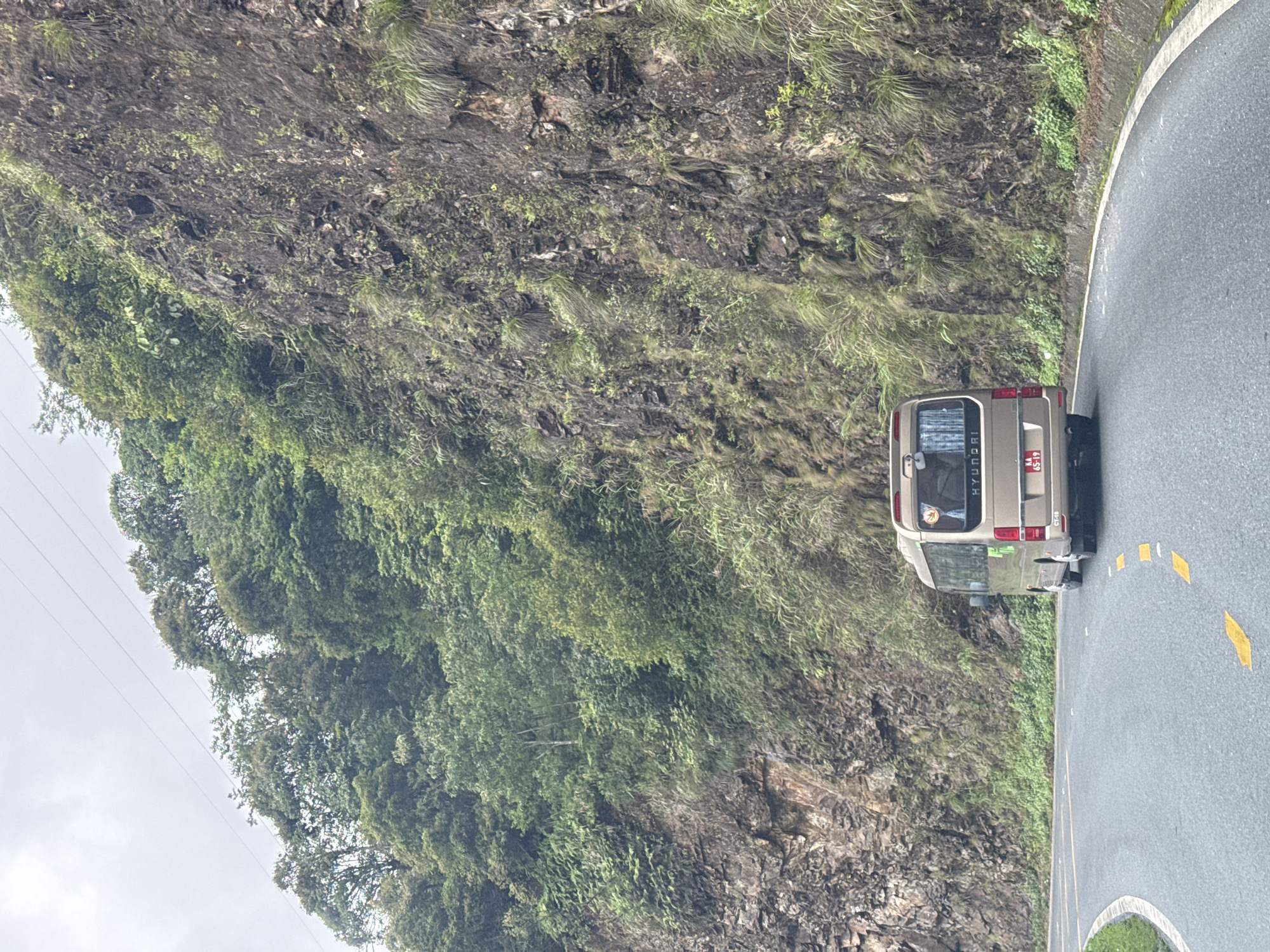 A tan Hyundai van with license plate BA 6559 is parked on the side of a paved road in Sa Pa Ward, Vietnam. The road curves sharply to the right, with a cliff face covered in vegetation rising sharply on the left. Yellow painted road markings are visible on the pavement. Power lines are present in the upper left.
