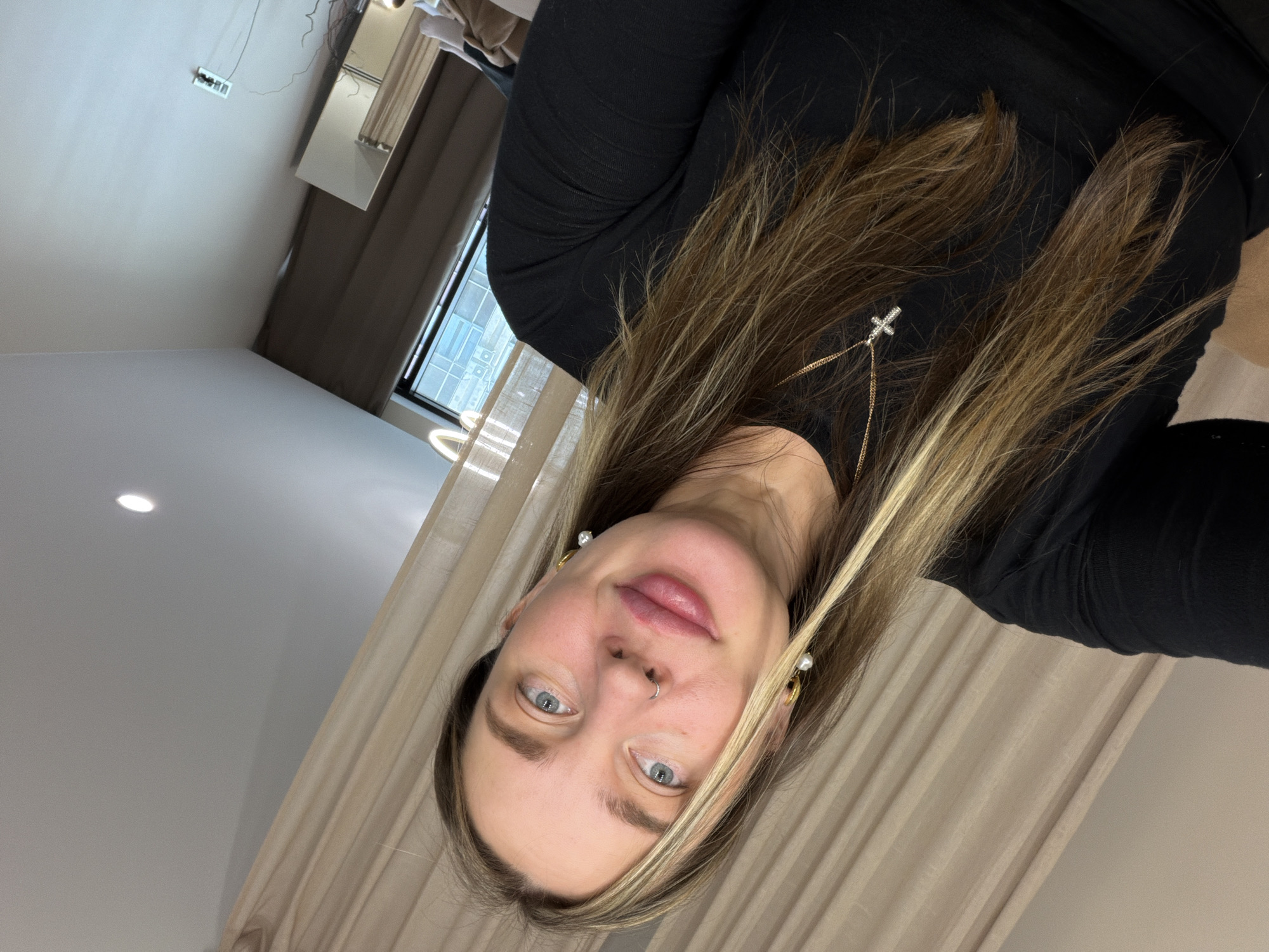 A woman with blue eyes, fair skin, and long brown hair with blonde highlights poses for a photo indoors. She has a nose piercing and is wearing a black long-sleeved shirt and gold earrings. A gold necklace with a cross pendant is visible in her hair. The background features a beige curtain, a window, and a ceiling with a light fixture. The location is possibly in Dolgoprudny, Russia.