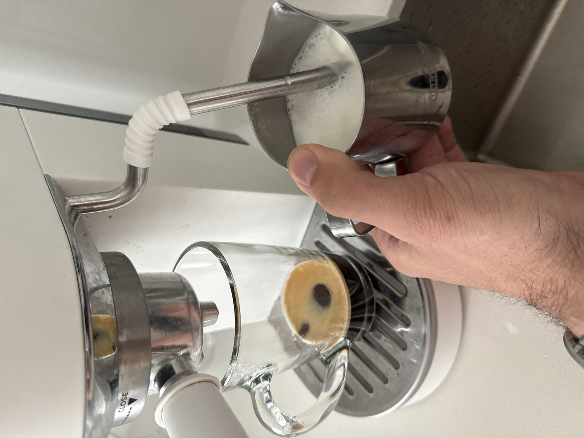A hand holds a silver pitcher and pours frothy milk into a clear glass mug containing a dark liquid, presumably coffee, on what appears to be an espresso machine. The machine is white and silver, with a stainless steel grate beneath the cup. The pitcher is marked with the word "DeLonghi". The setting is indoors and the location details of Perm, Russia are not evident in the image.